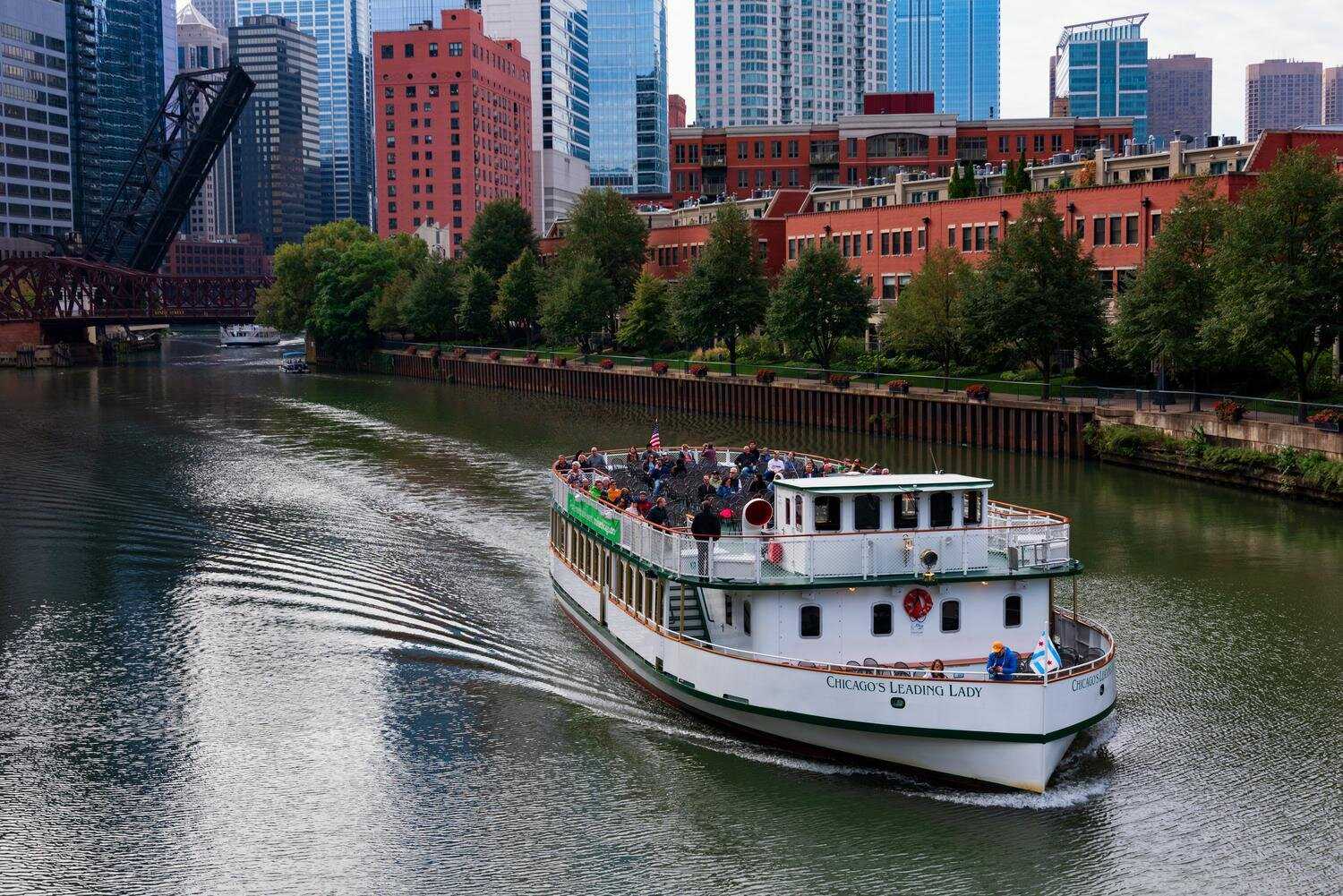 A large boat cruises down the river, with tall buildings on either side.