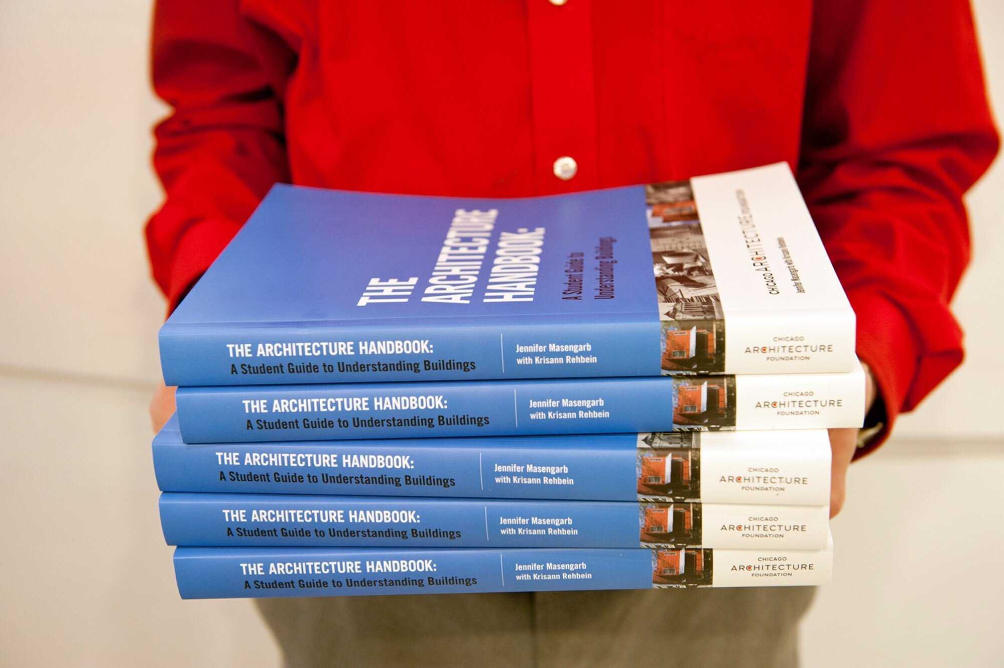 Close up of a person in a red shirt holding a stack of books titled The Architecture Handbook