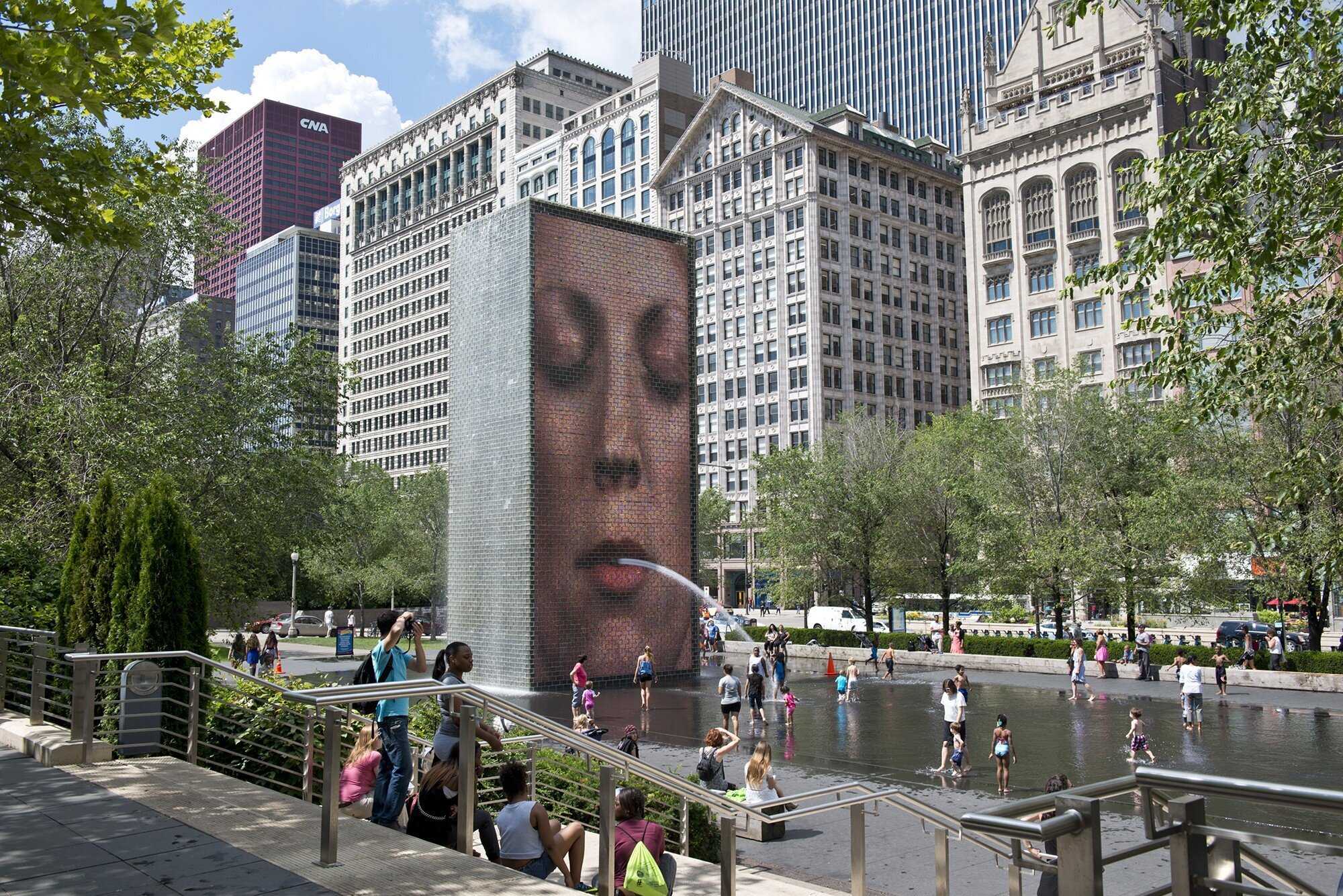 Image of a large tall rectangular structure that has a face on the front with water coming out of the mouth of the person. Behind the fountain there are buildings and people are standing in front of it. 