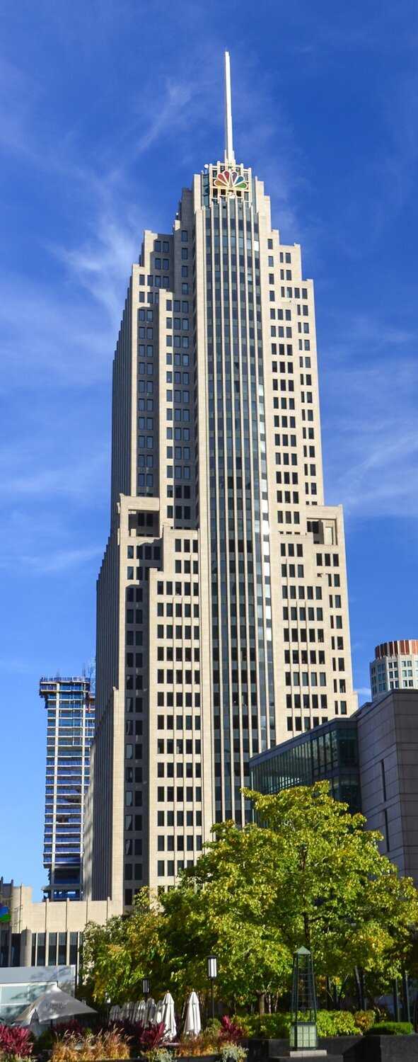 Photo of a tall white building with a spire on top and an NBC logo on the facade with rainbow peacock feathers