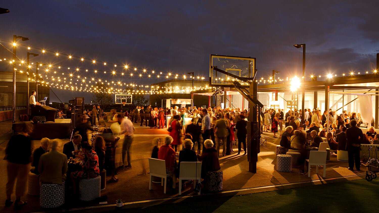 A night event outside with fairy lights above many tables with people seated and enjoying themselves