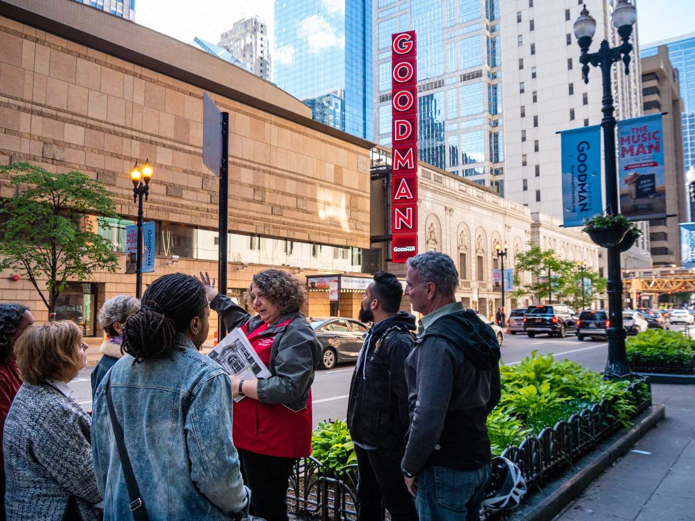 Woman in red jacket points across the street to a theatre with a red sign utside that says Goodman. She is speaking to a tour group