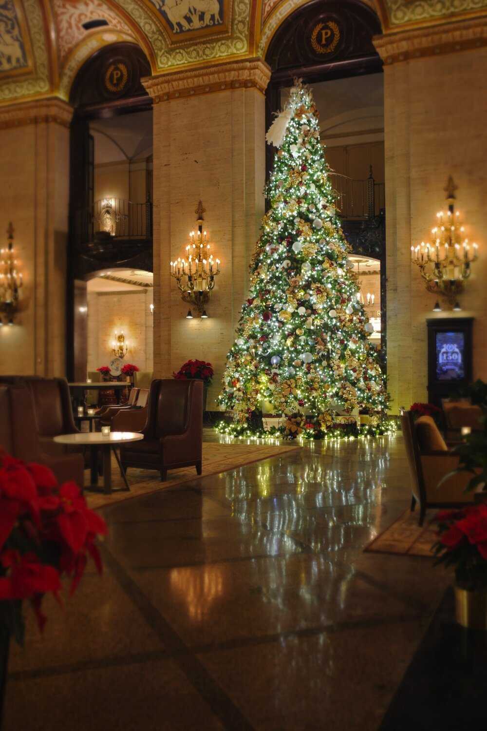 Picture of the interior of a hotel lobby which contains a decorated Christmas tree, furniture, golden light fixtures, and a painted ceiling