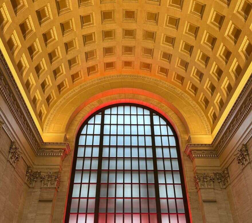 Ornate arched ceiling with squares carved into it above a large window framed by columns and lit with a red light