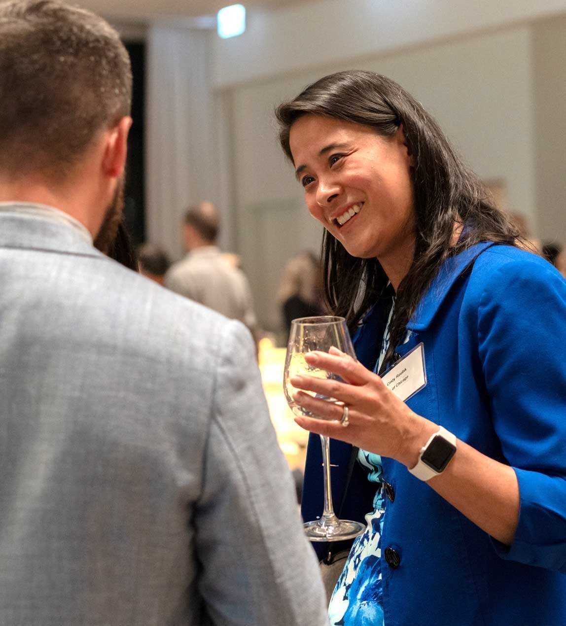 Asian woman in a blue top holding a glass of wine and smiling and speaking with a man with a grey suit at an event.