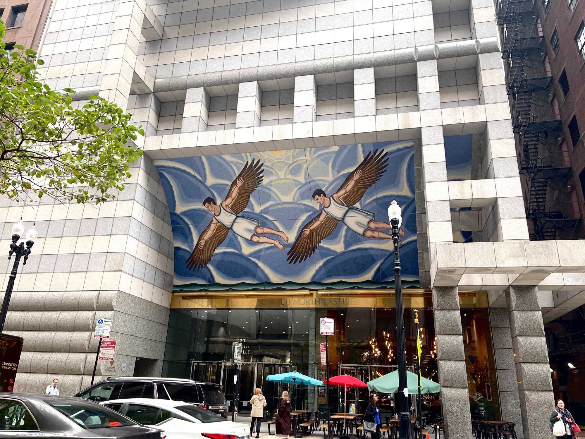 Image of the front of a building with a mural of two men with wings flying in front of a blue background in front of a door.