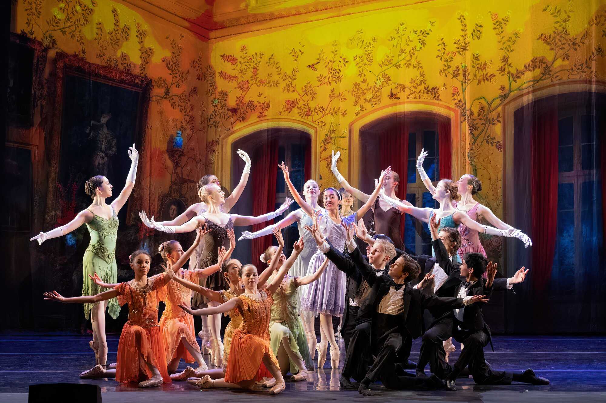 Image of several ballet dancers in formal attire standing together with their arms outstretched on a stage with a yellow backdrop that is covered in paintings of plants and birds.