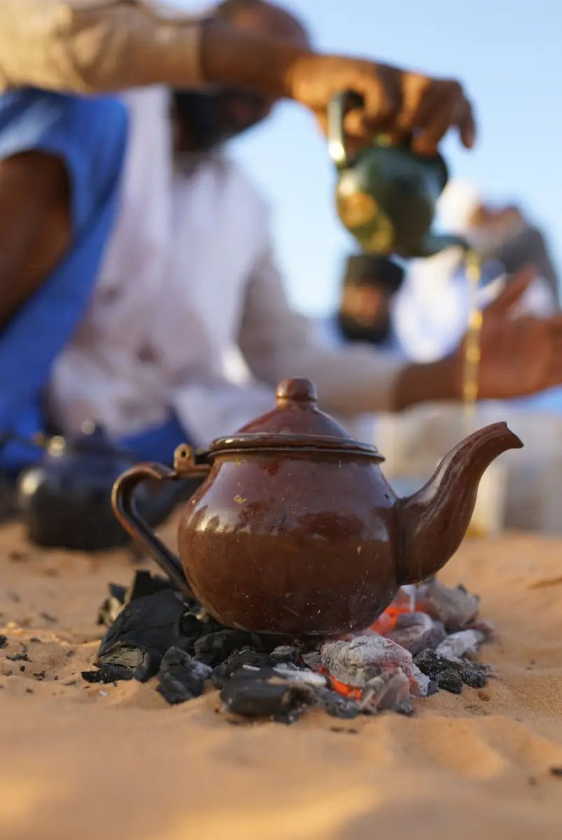 Tea being brewed in a metal teapot set on embers in the sand, poured from a height into glasses