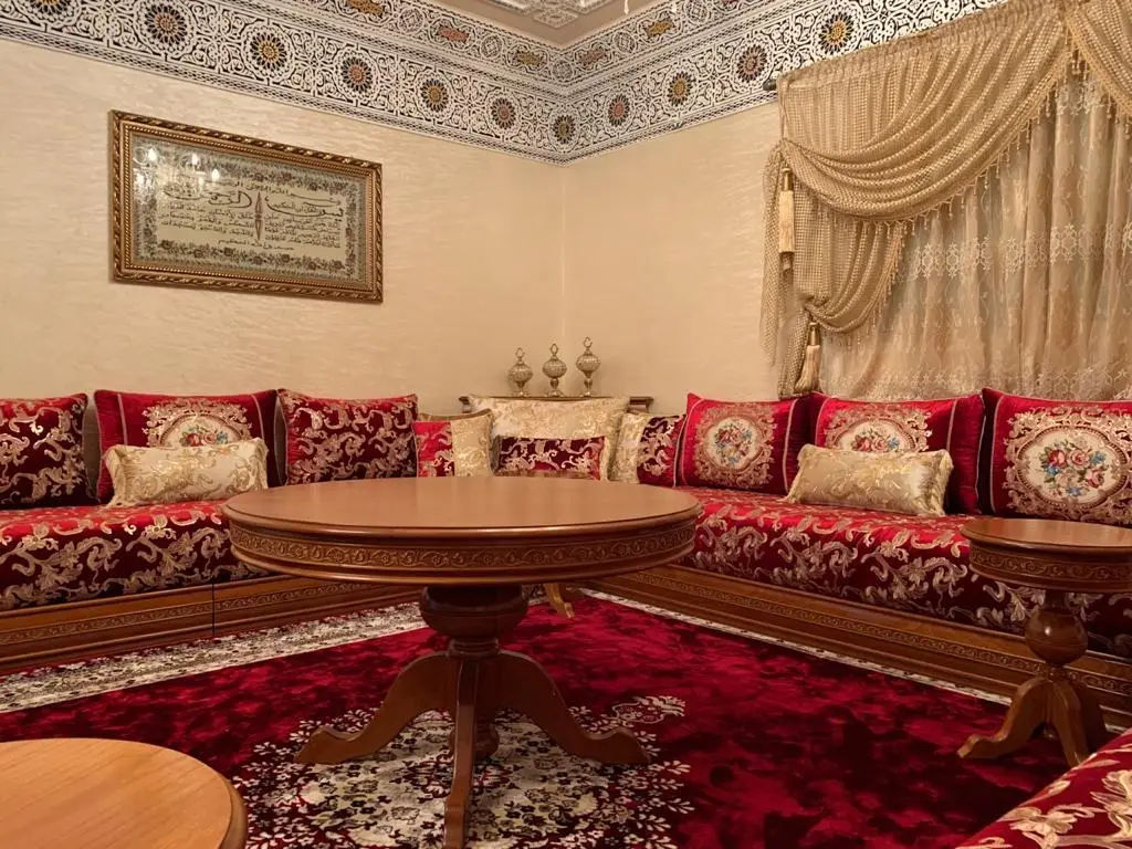 Salon marocain traditionnel avec sedari sur socle en bois, housses rouges décorées et table ronde