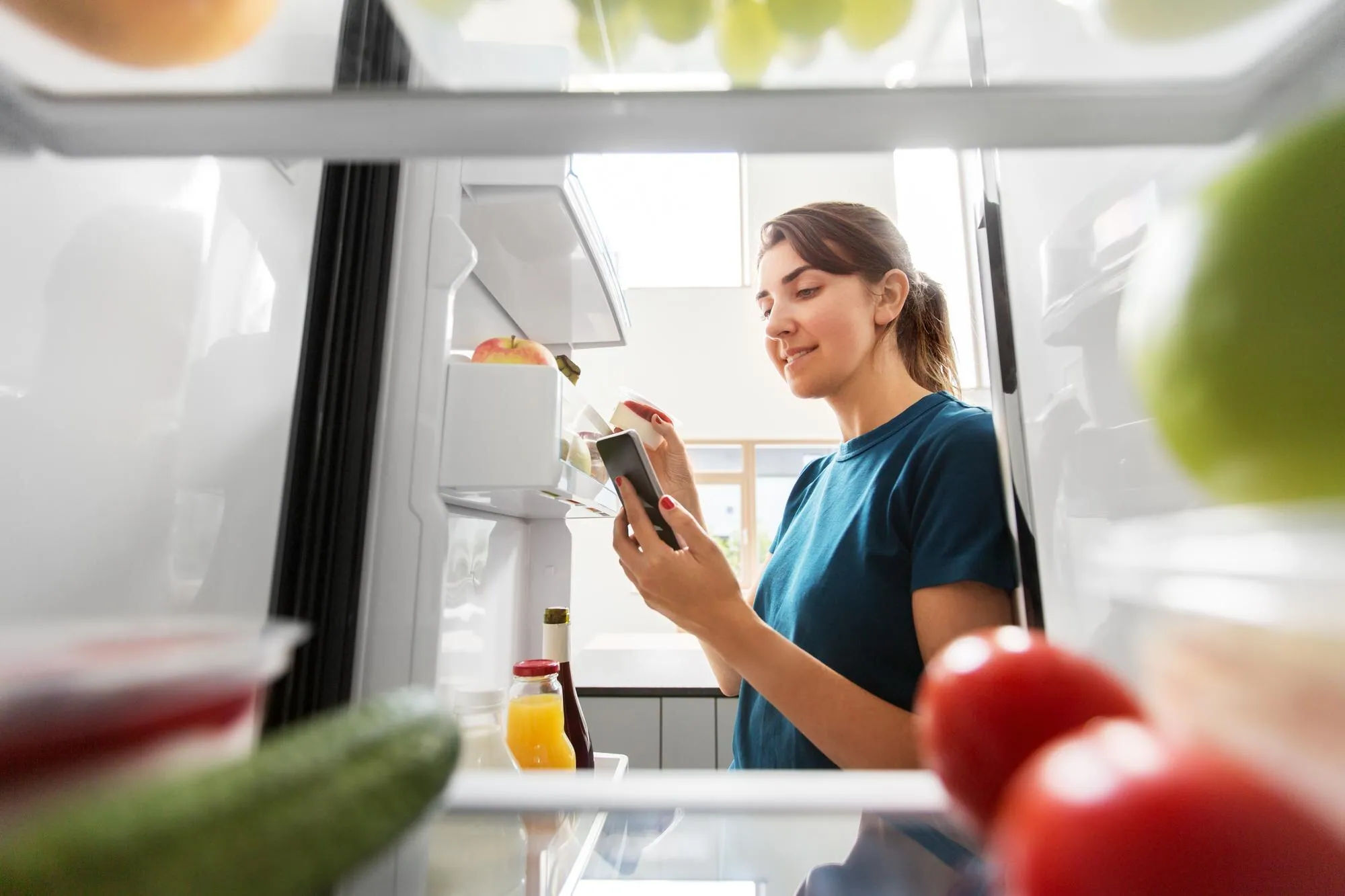 Женщина проверяет продукты в холодильнике и составляет список в телефоне