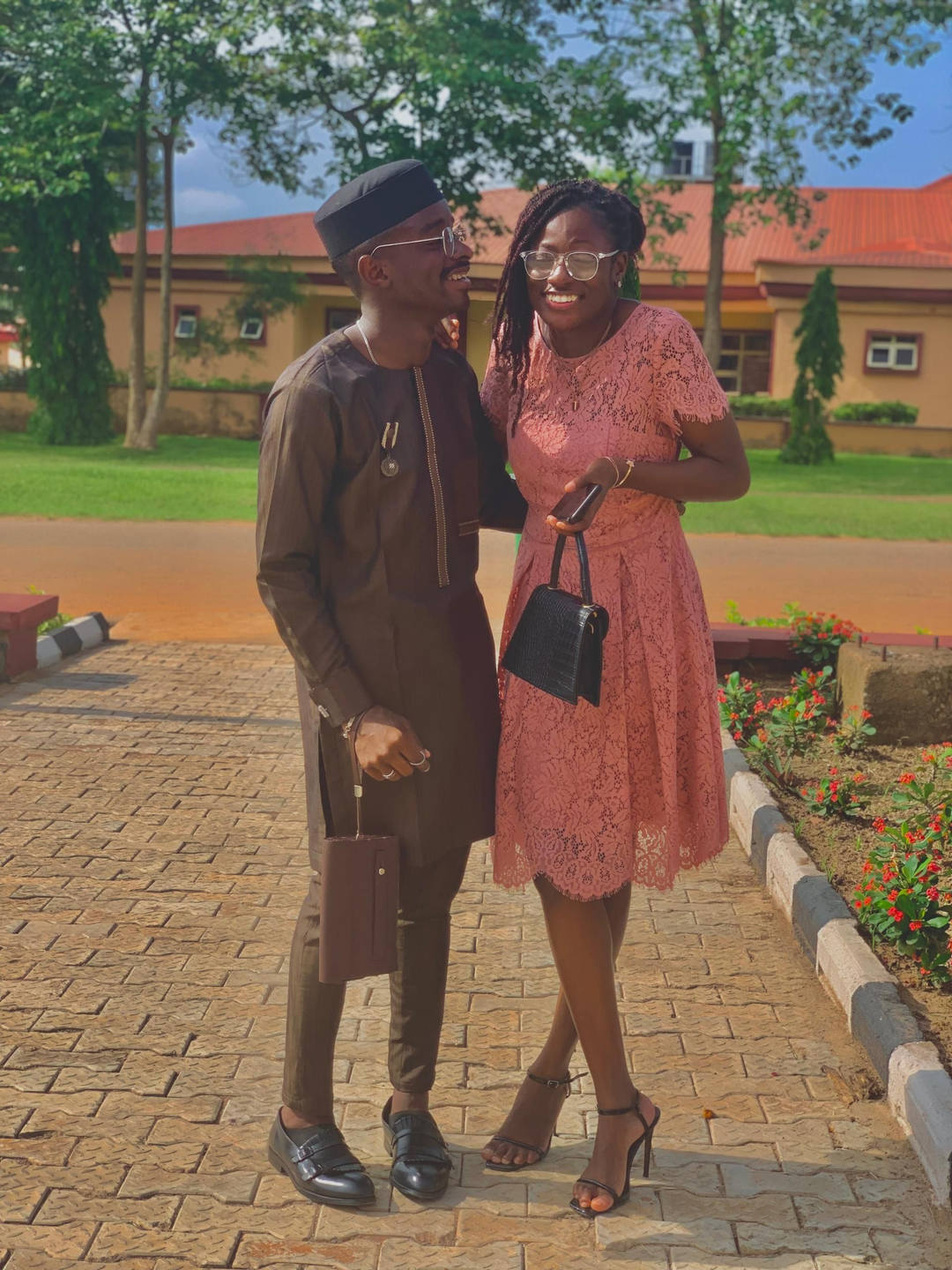 Mary in a pink lace dress and Paul in a dark kaftan and cap, laughing together on a campus path.