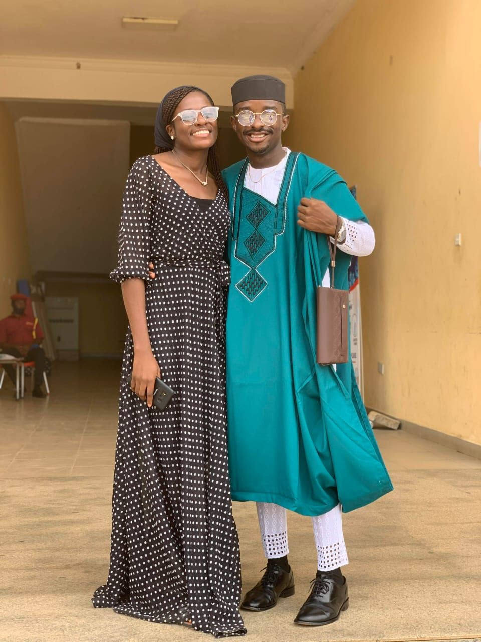 Mary in a black polka-dot maxi dress beside Paul in a teal agbada, outside a church hall.