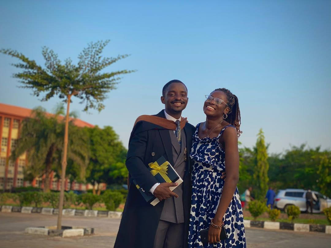 Paul in a graduation gown holding a wrapped gift, Mary beside him on a sunlit campus road.