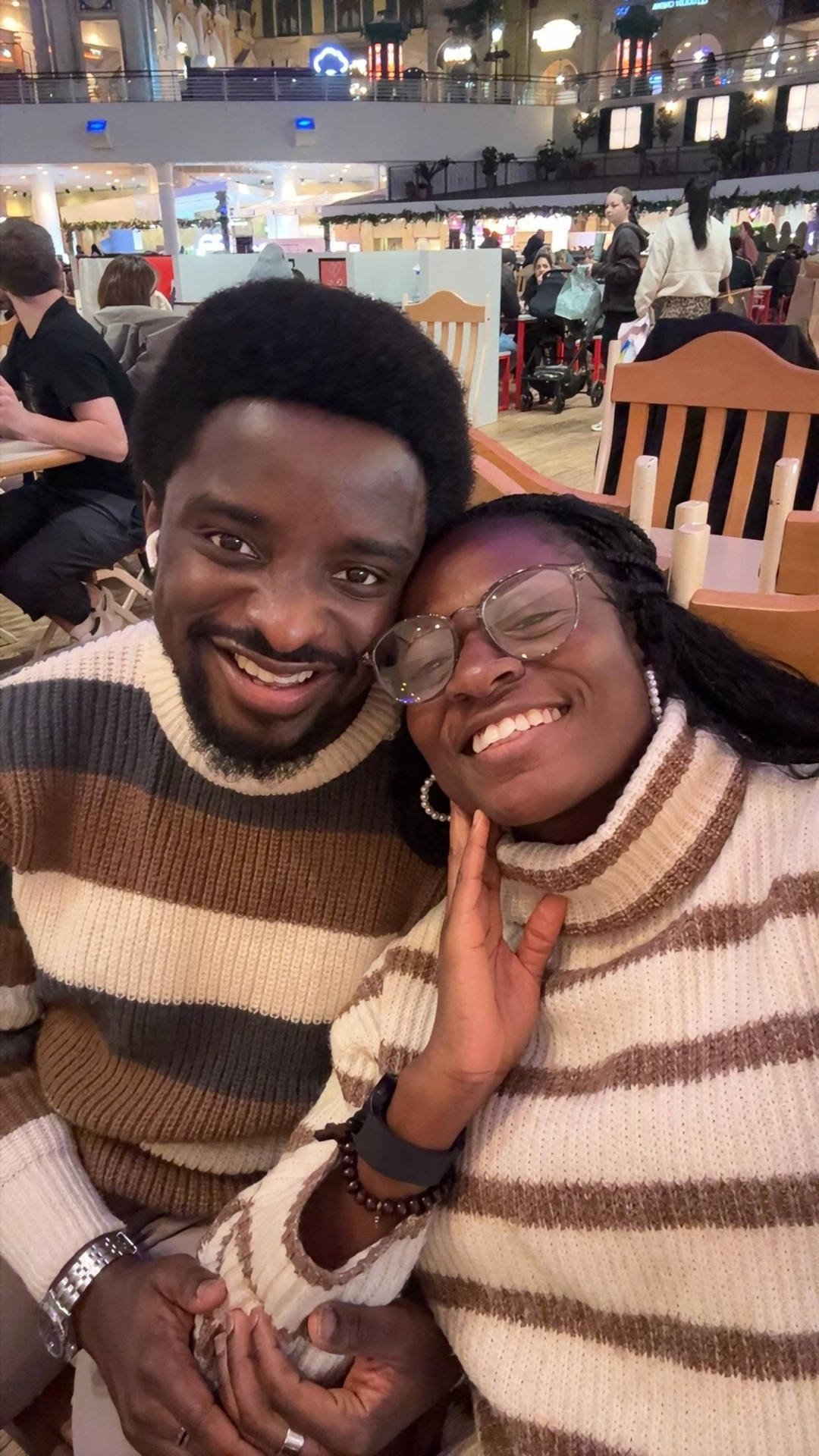 Mary and Paul in striped knitwear, heads together at a table in a busy shopping centre.