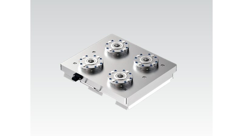 Rectangular silver zero-point base plate with four round receivers, each with a center bore and a ring of blue screws, and a small dark fitting on the left edge