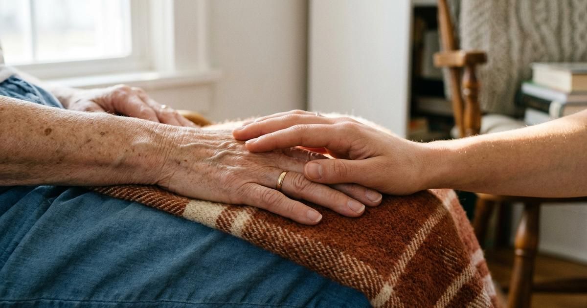 A hospital bed set up in a familiar living room with a lamp, a folded quilt and a chair pulled close beside it for a family member to sit.
