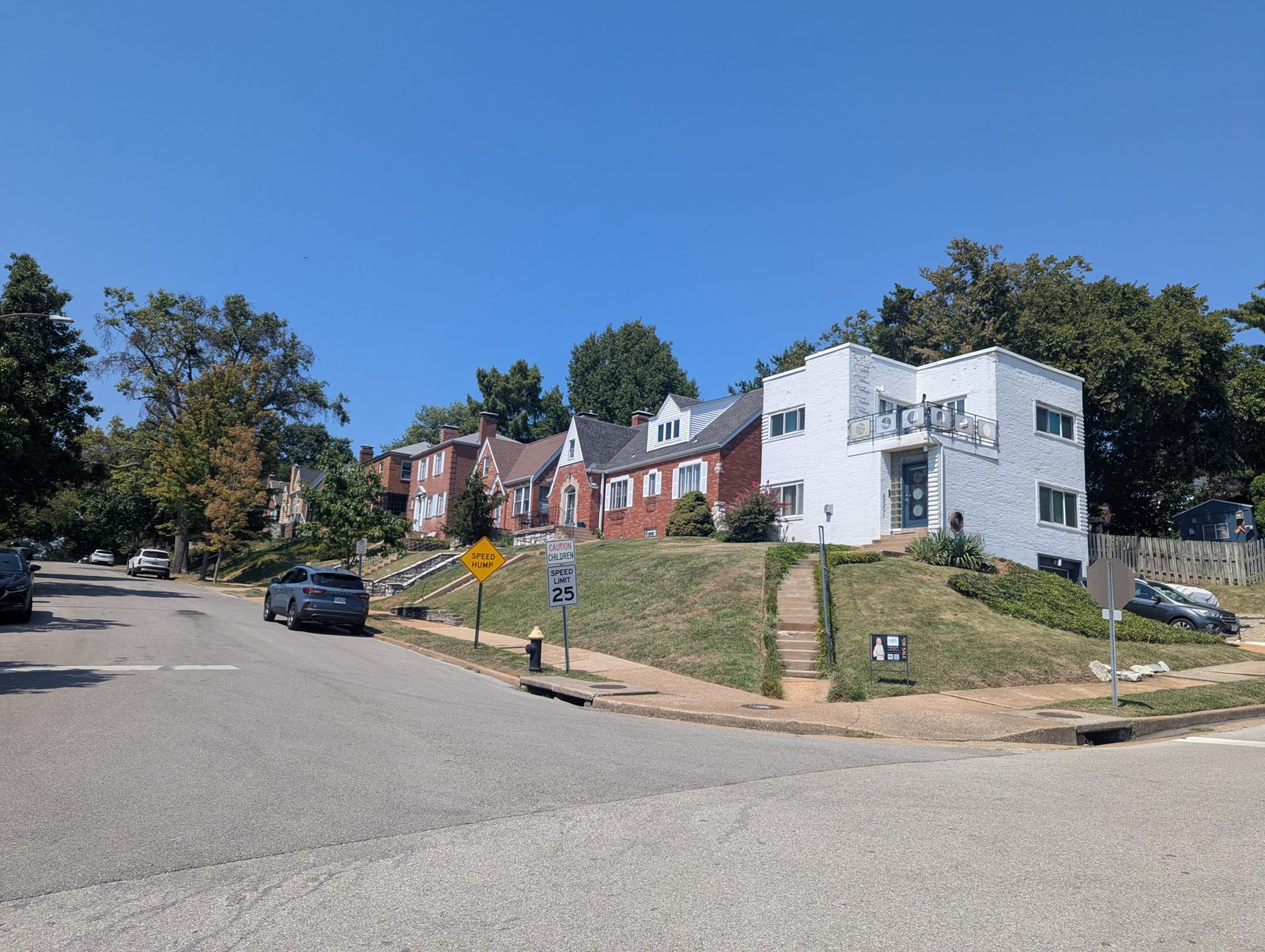 A hillside block of red brick homes in south St. Louis with a white Art Deco corner house and a for-sale sign at the steps