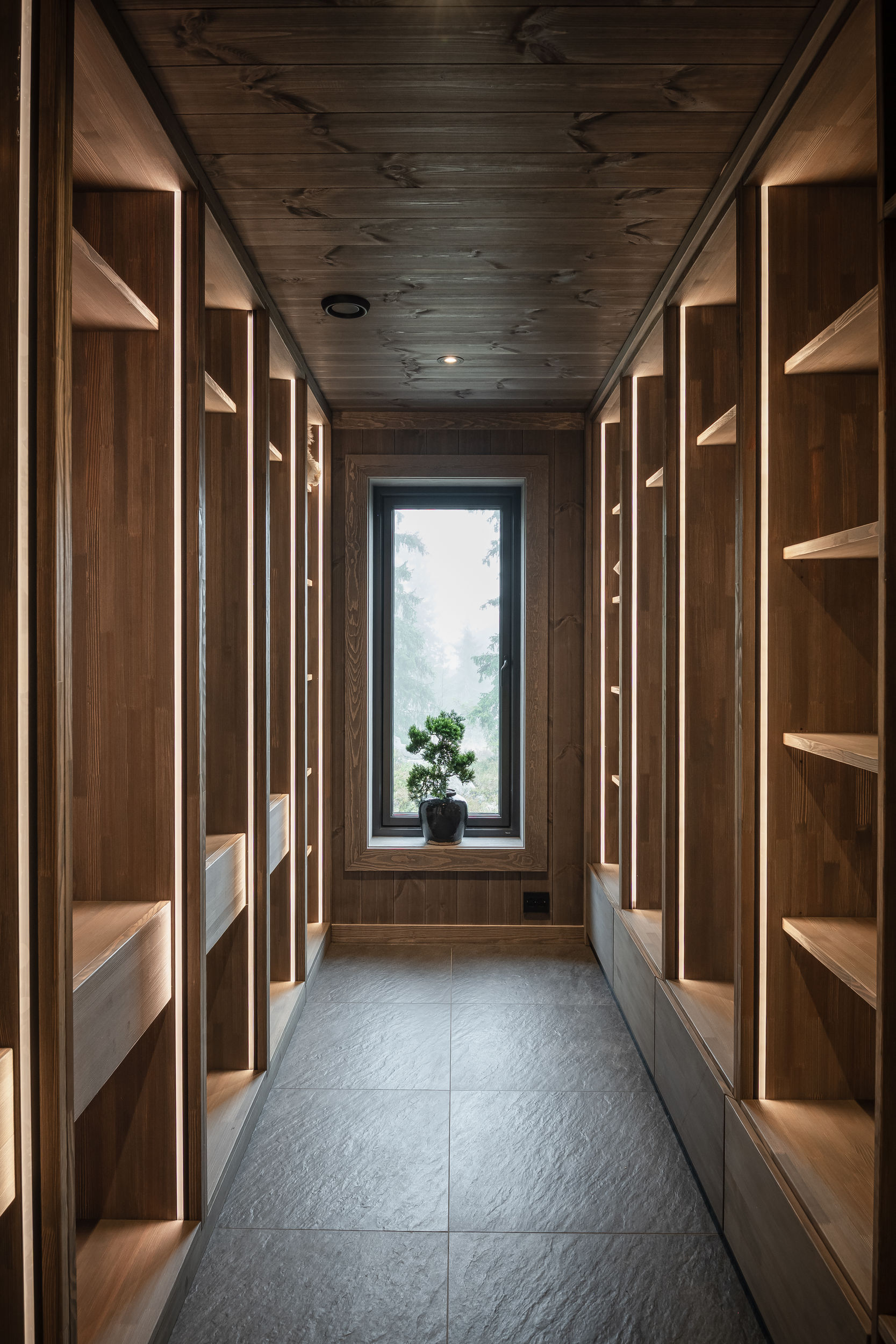 Plassbygget walk-in-garderobe med åpne trehyller, skuffer og integrert belysning langs en smal gang.