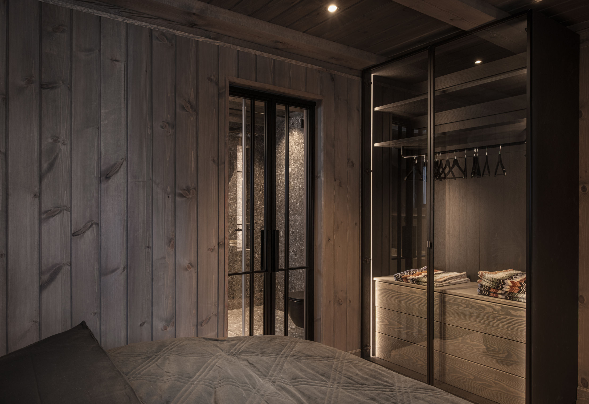 Plassbygget garderobe med glassfronter, skuffer og klesstang i et mørkt hyttepreget soverom.