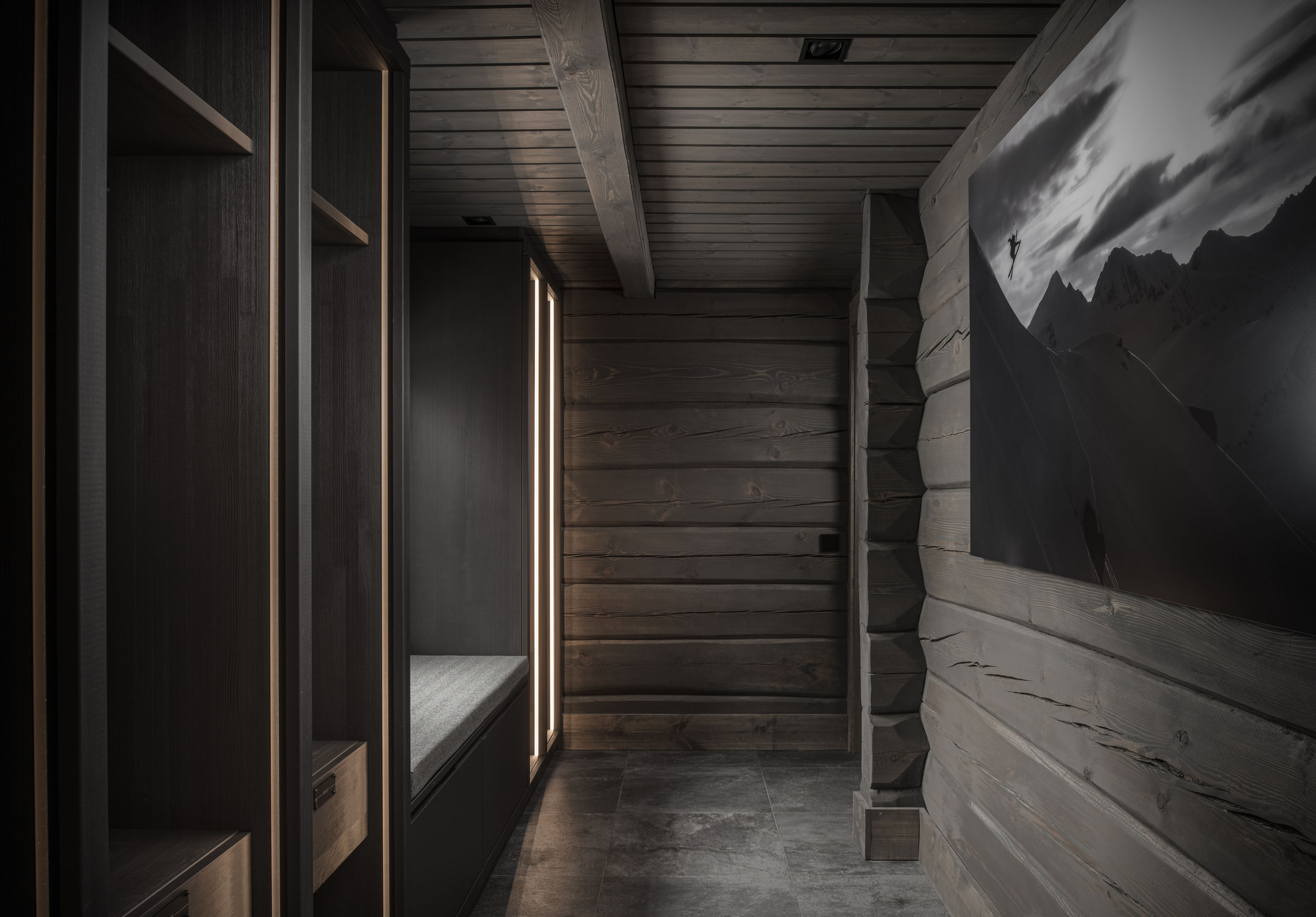 Plassbygget garderobe med sittebenk, åpne hyller og lysfelt i mørk hyttegang.