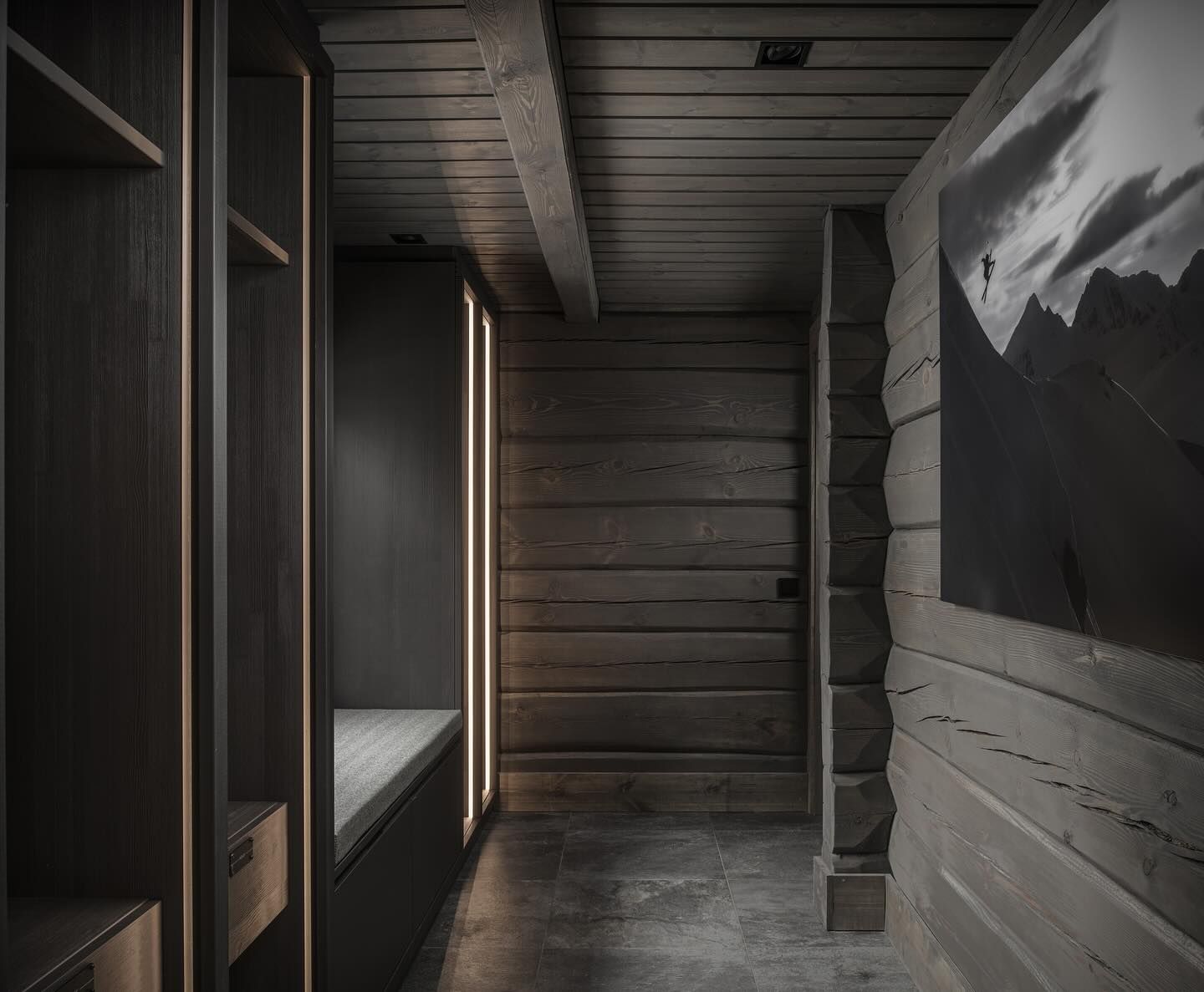 Mørk plassbygget entrégarderobe med sittebenk, åpne hyller og vertikale lysfelt i hyttegang.
