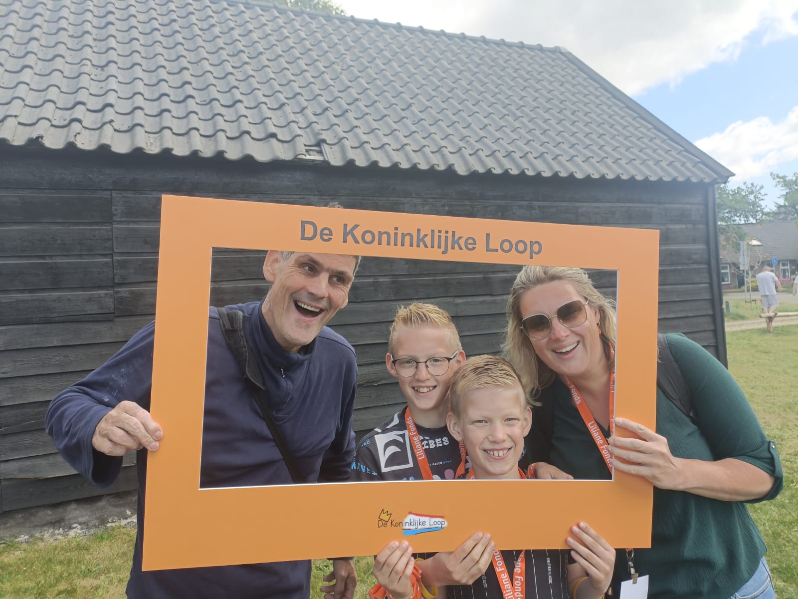 Gezin met kinderen houdt een oranje frame met 'De Koninklijke Loop' vast voor een houten gebouw in Apeldoorn