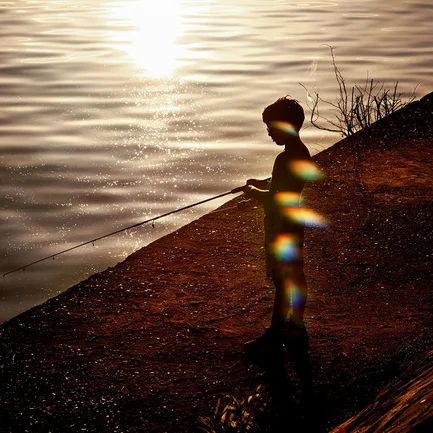 A photograph of a boy fishing on the shore of a lake.