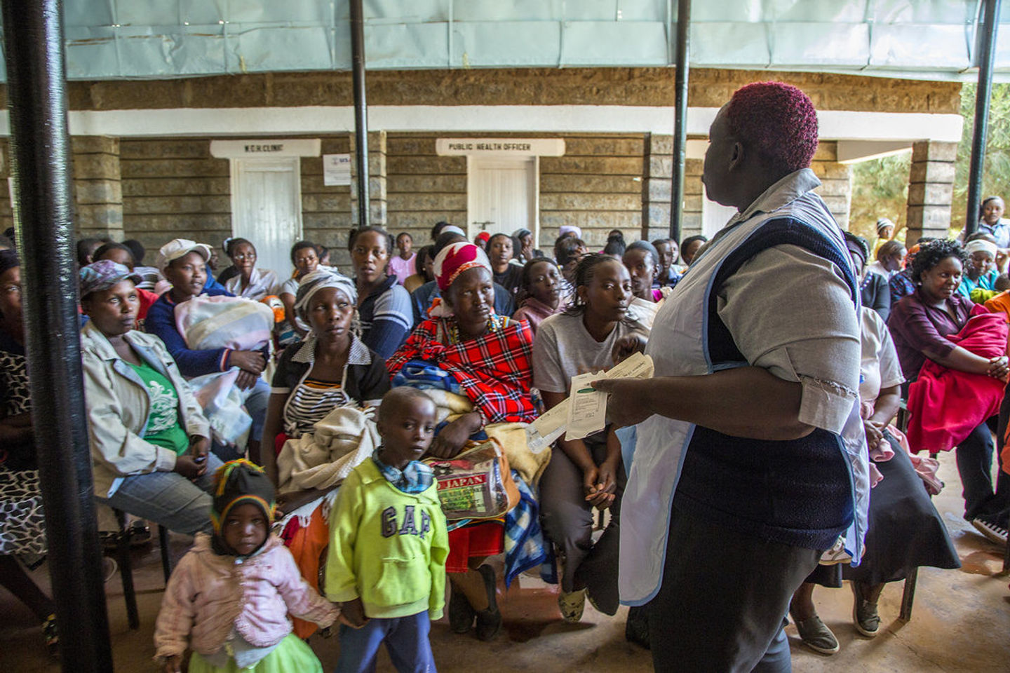 A health worker addressing a seated community gathering outdoors.