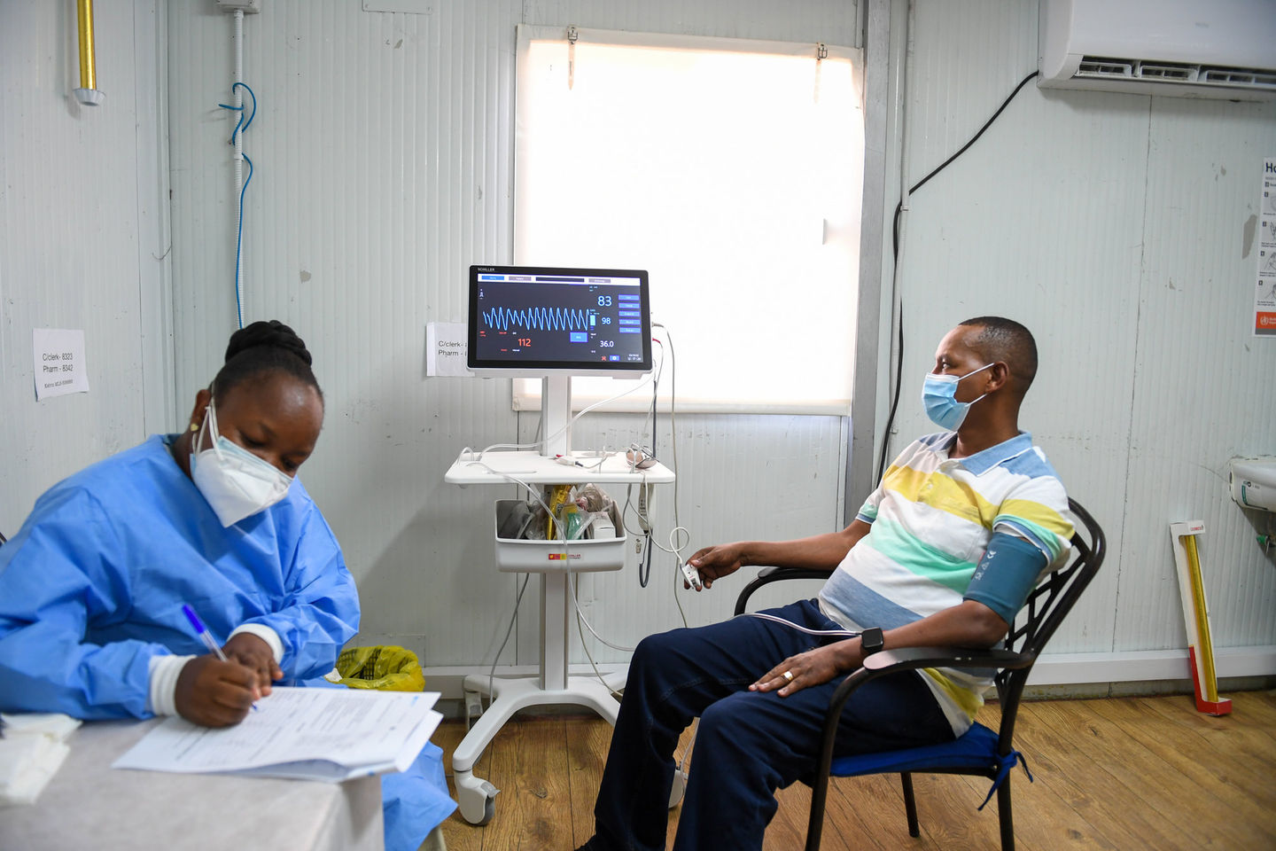 A health worker in protective equipment attending to a seated patient in a clinic.