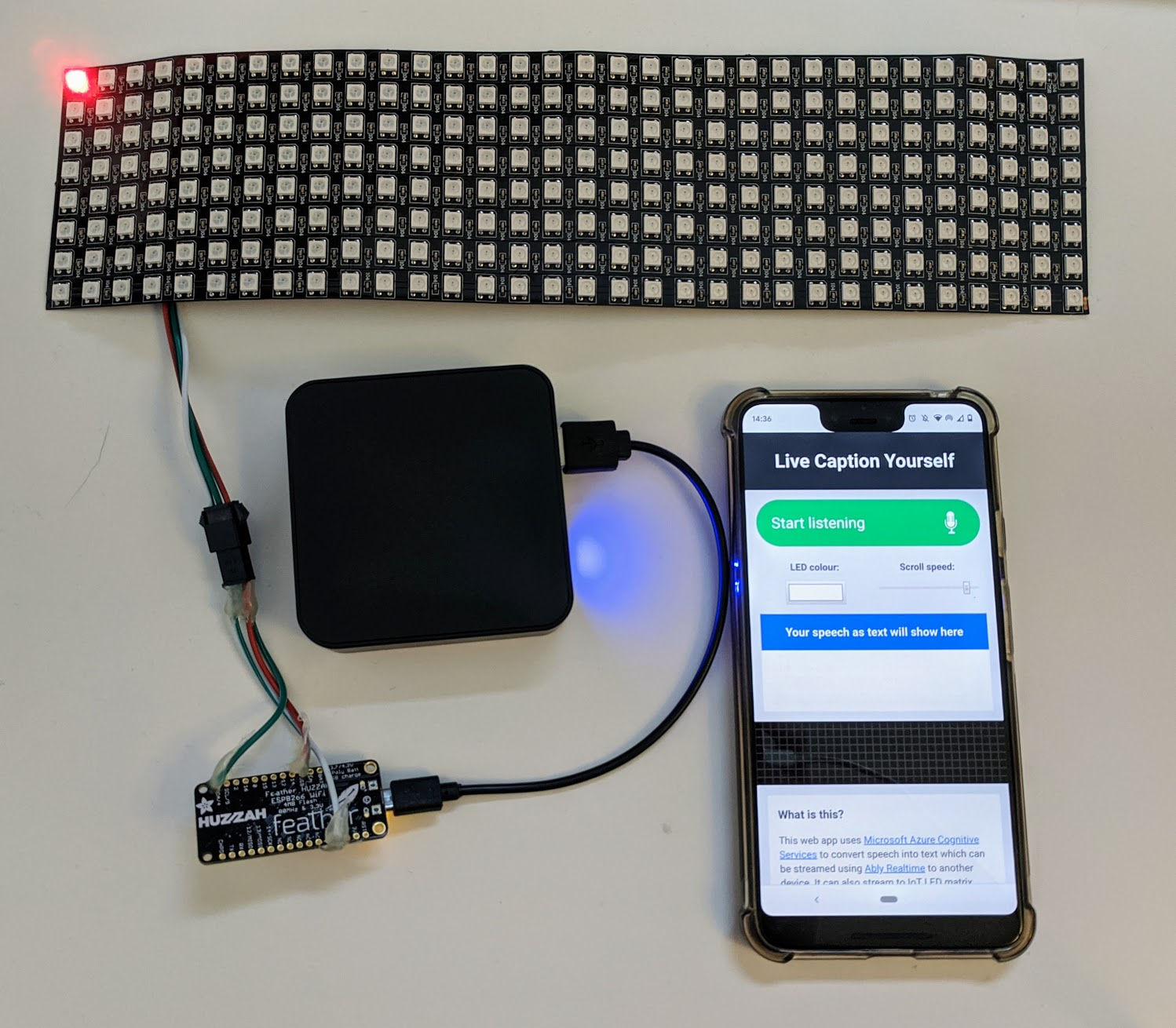 A flexible display attached to an Adafruit Feather Huzzah connected to a USB battery and a phone displaying the app