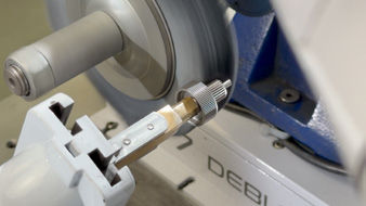 A pinion shaft held by custom gripper fingers approaches the finishing wheel.