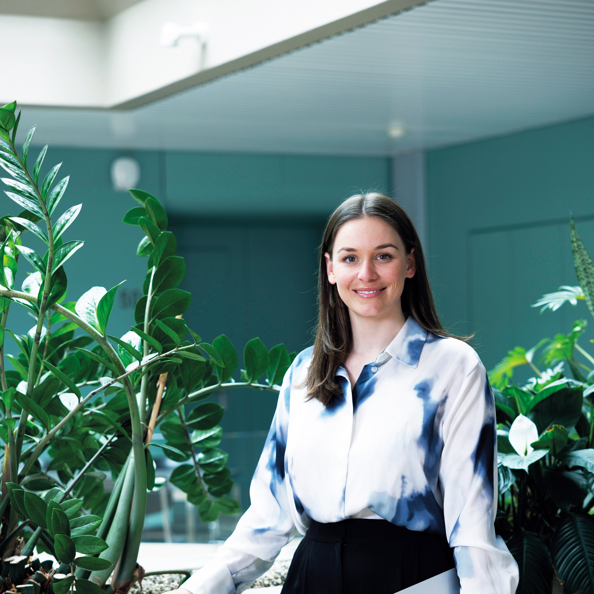 Eine junge Frau steht in der Akademie St.Gallen vor grünen Pflanzen und hält ein Notizbuch in der linken Hand.