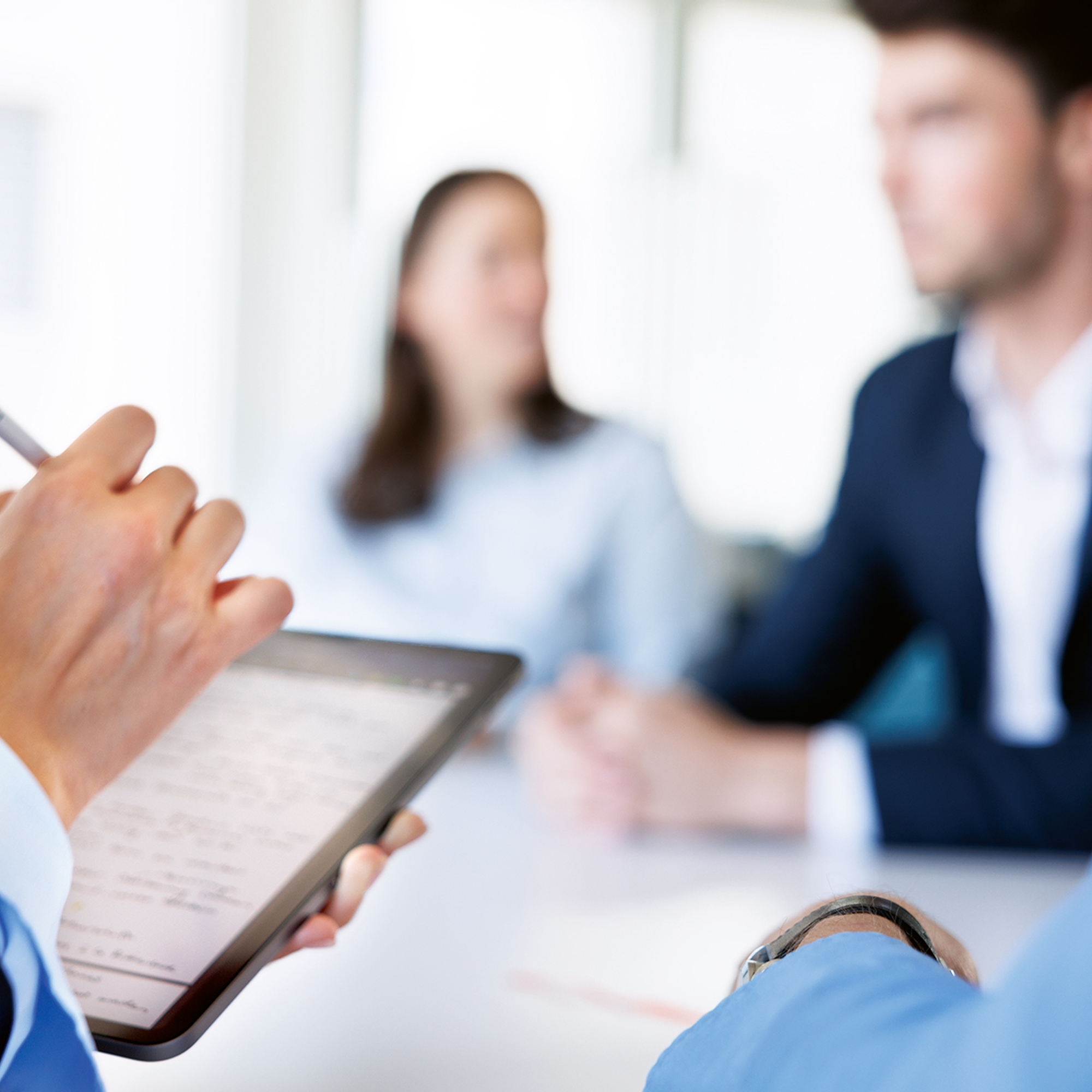 Ein IPad steht im Vordergrund, eine Hand schreibt etwas aufs IPad. Verschwommen im Hintergrund sieht man wie 2 Personen ein Meeting halten.