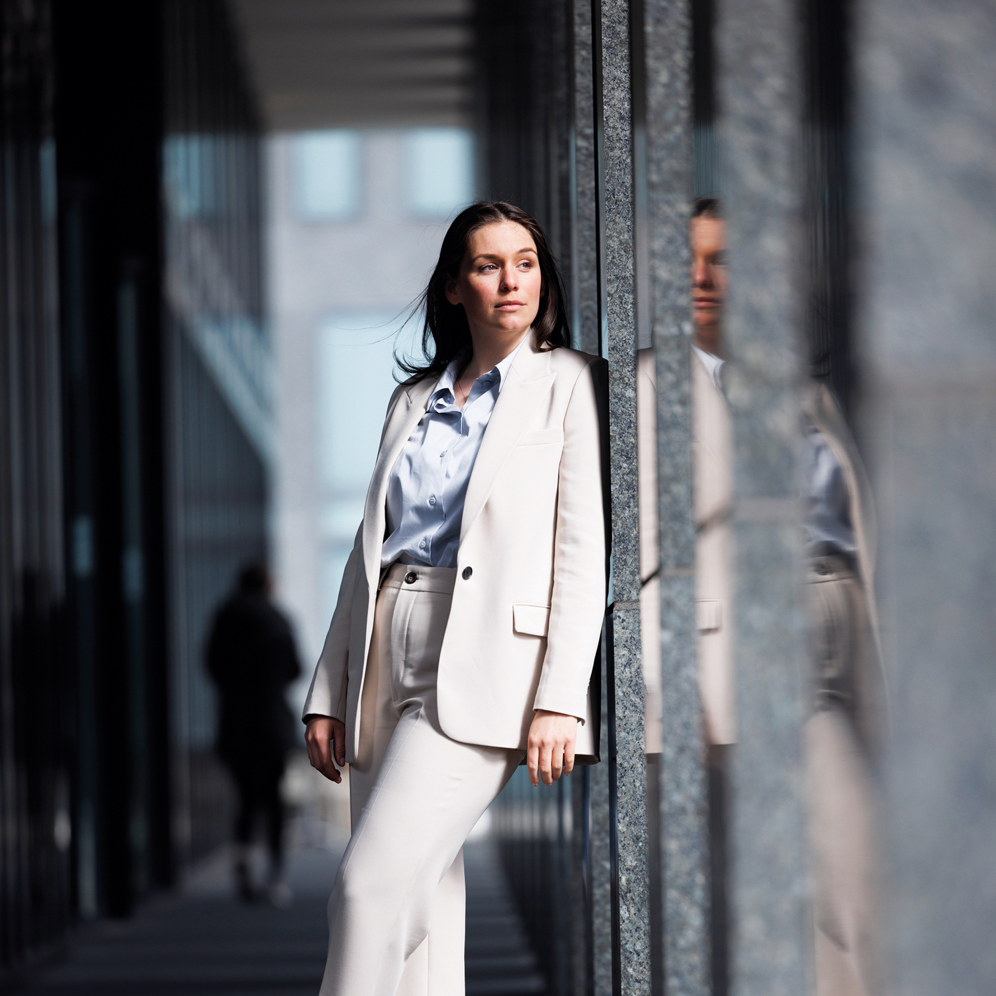 Eine Geschäftsfrau in weissem Anzug lehnt an der Aussenwand Ihres modernen Unternehmens