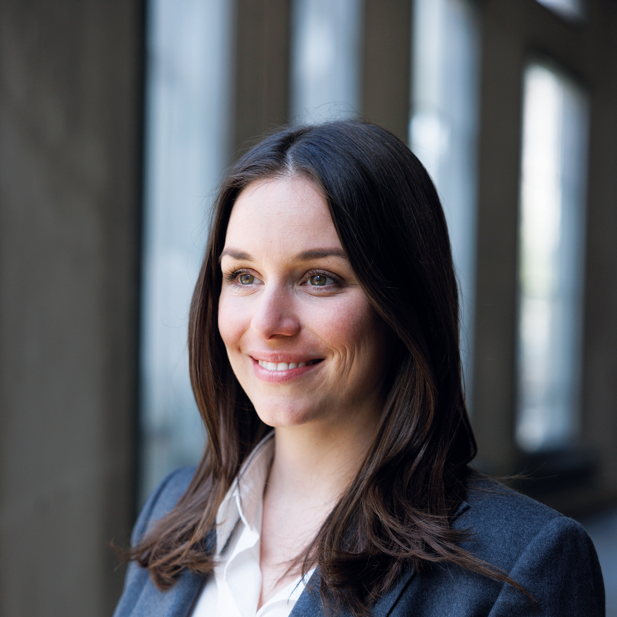Eine Businessfrau mit blauem Anzug uns schwarzem Haar schaut aus dem Fenster Ihres Bürogebäudes.