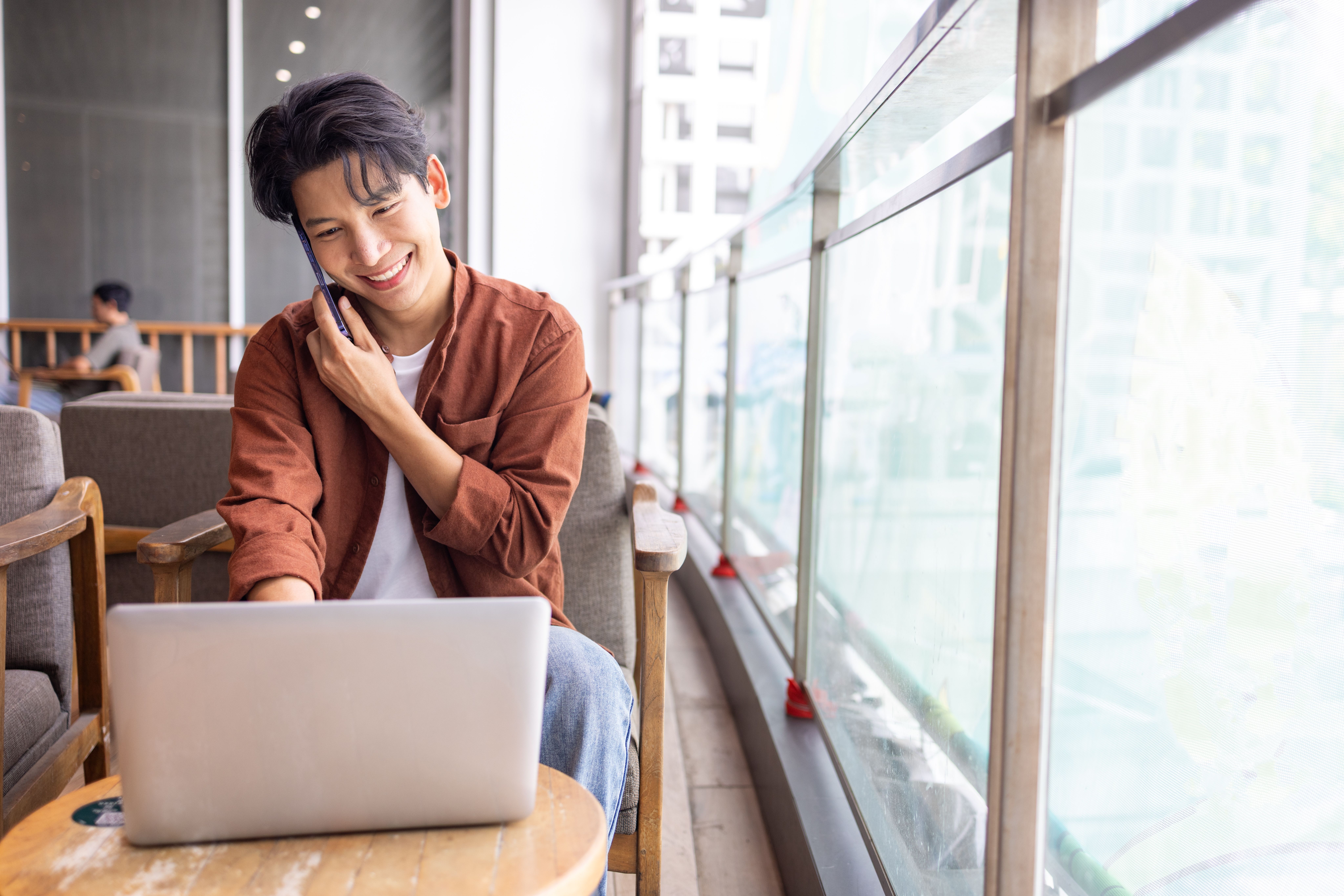 A young man sitting in a café, smiling while using a laptop and talking on a phone