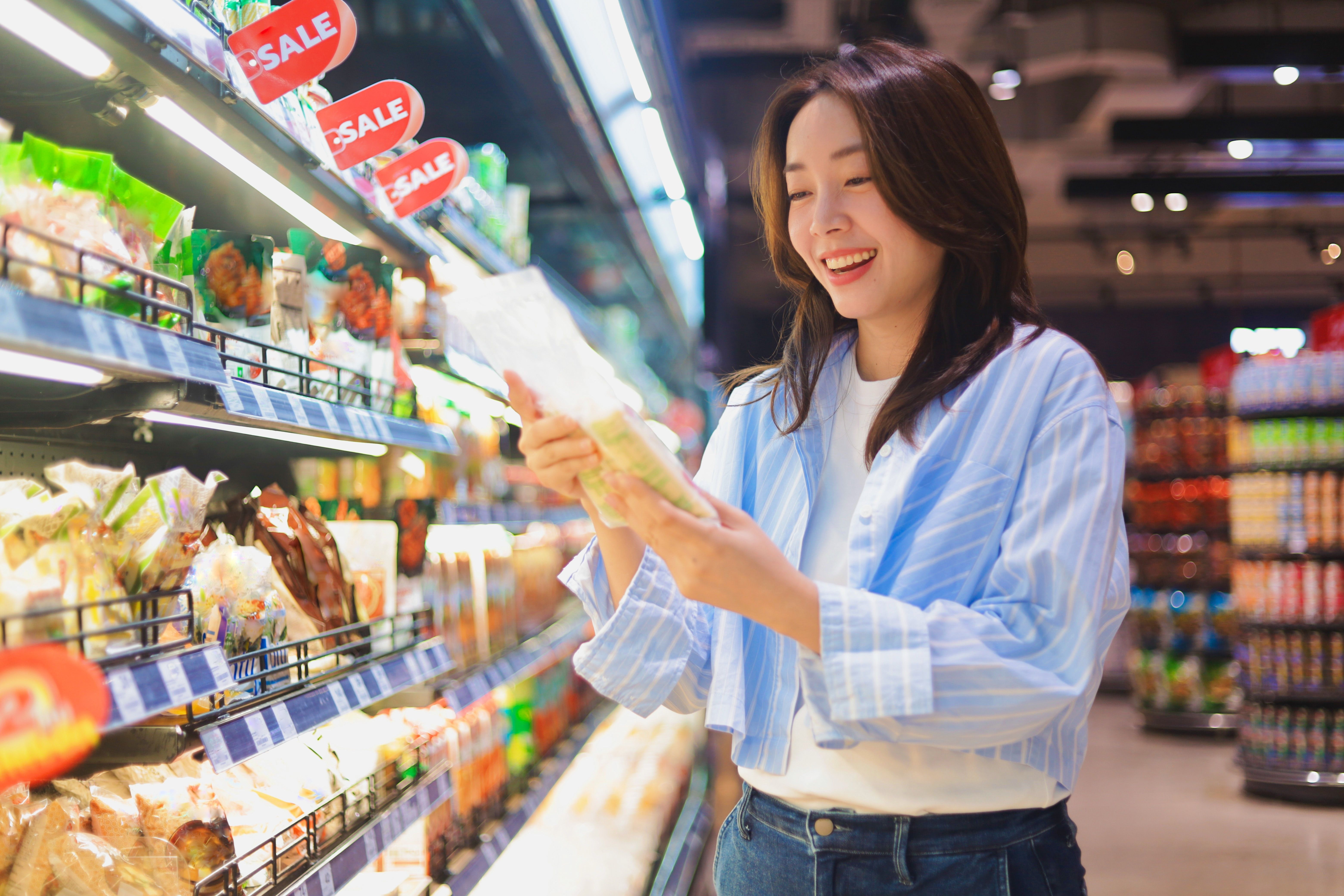 A woman smiling while shopping in a grocery store, examining a product on the shelves filled with various goods