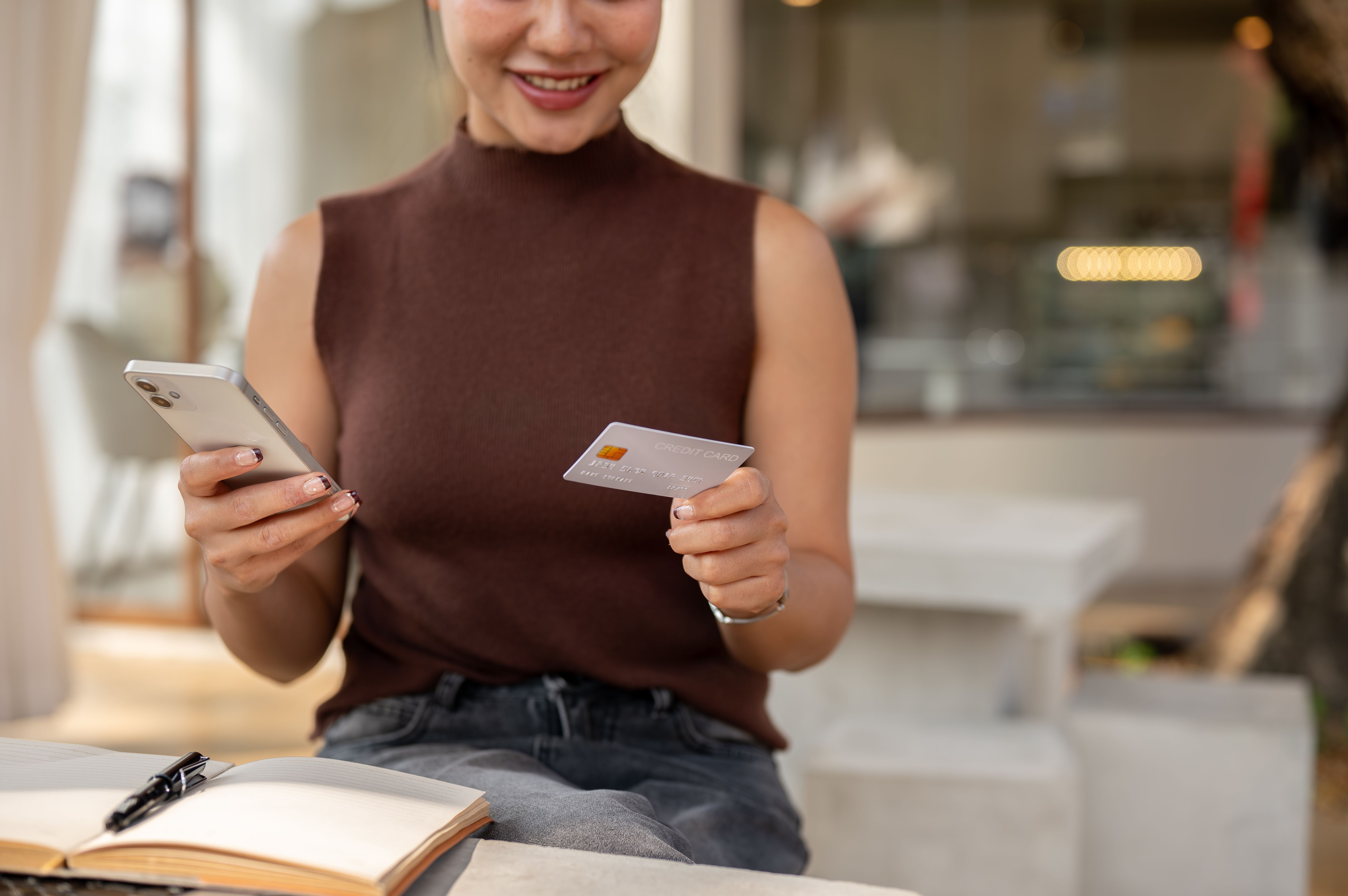 A woman sitting at a table holding a smartphone in one hand and a credit card in the other, smiling