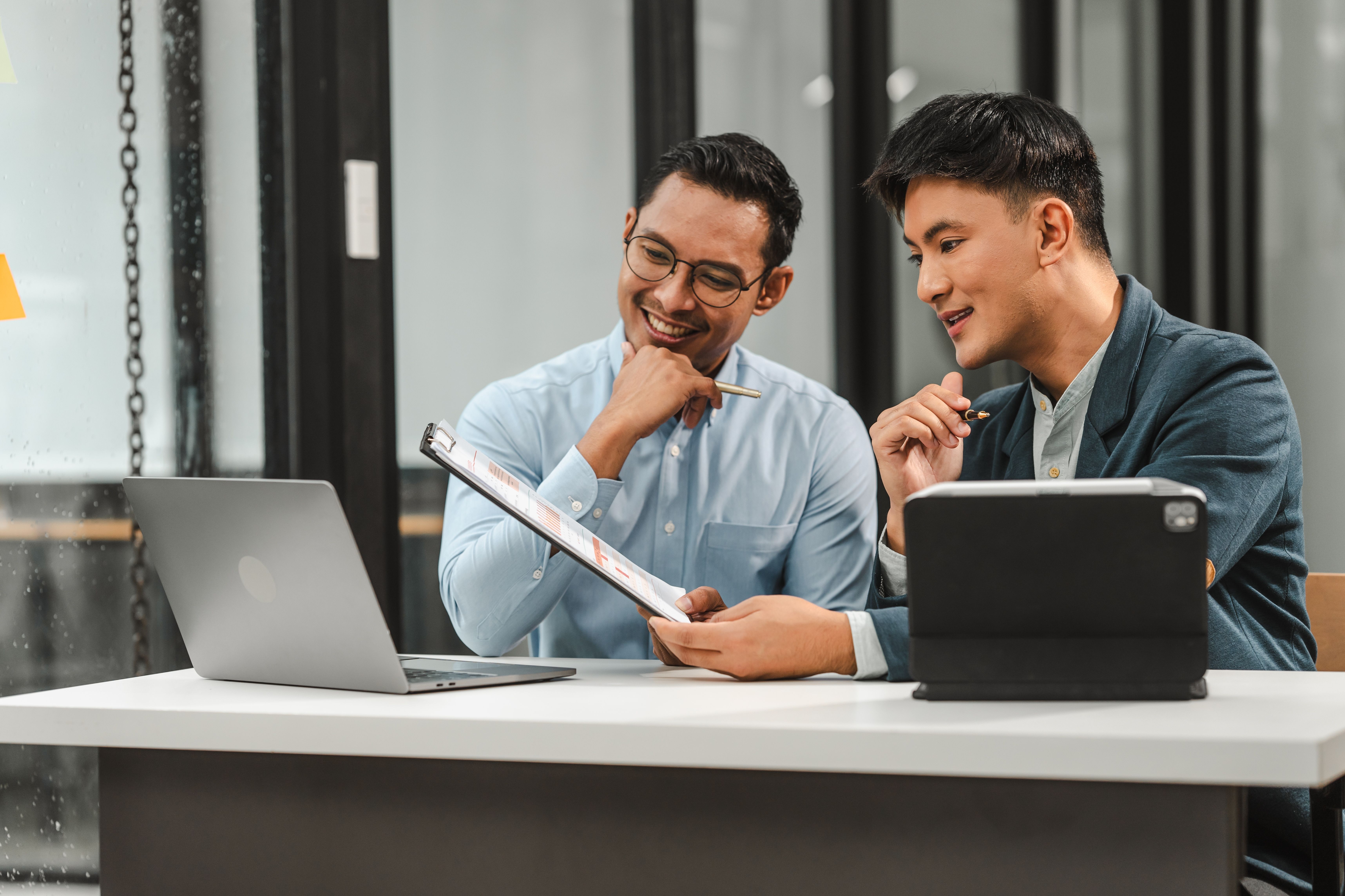 Two men sitting at a table, discussing something on a laptop and a tablet