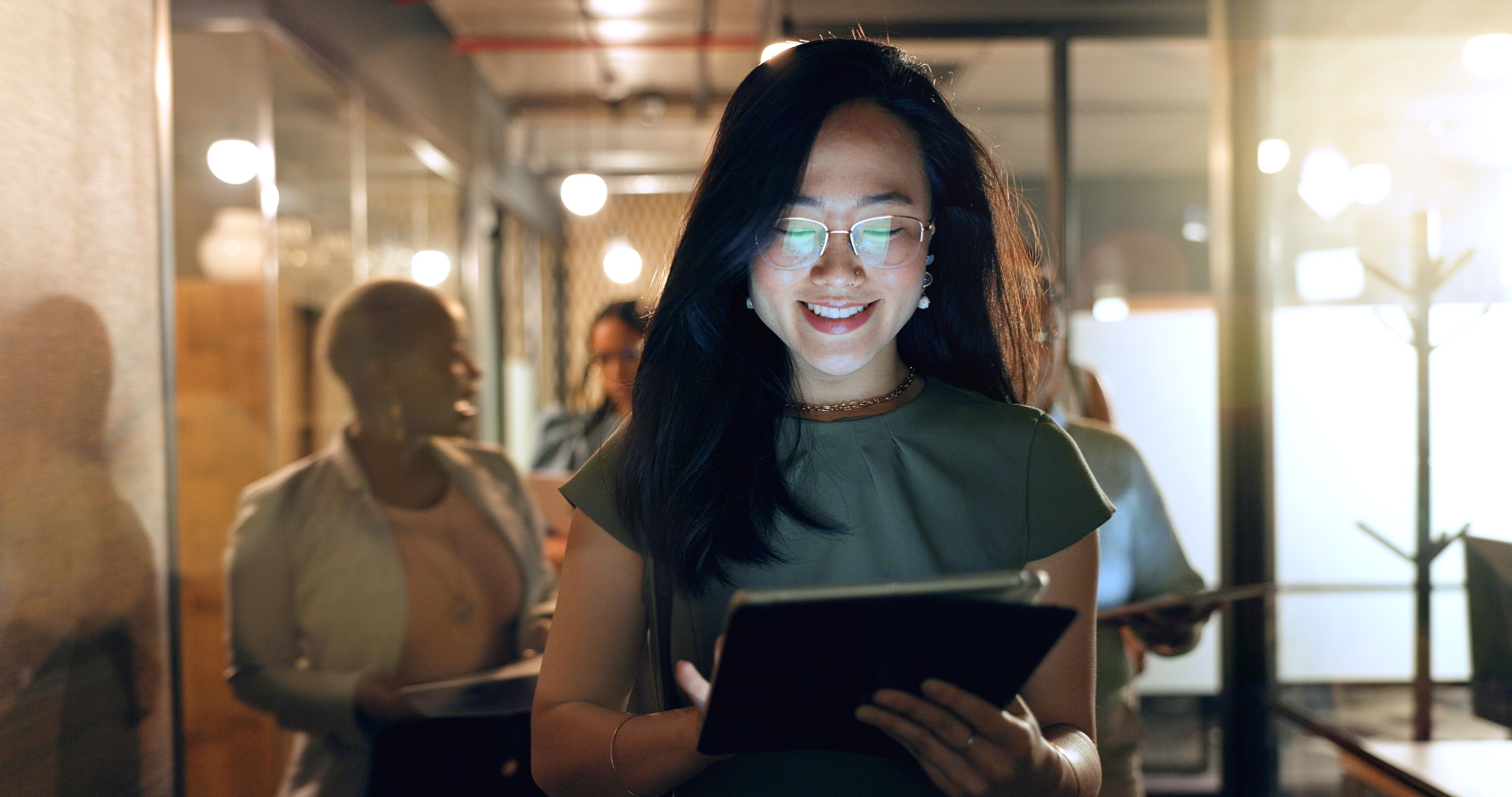 A woman with glasses is smiling while holding a tablet in a well-lit corridor, surrounded by people