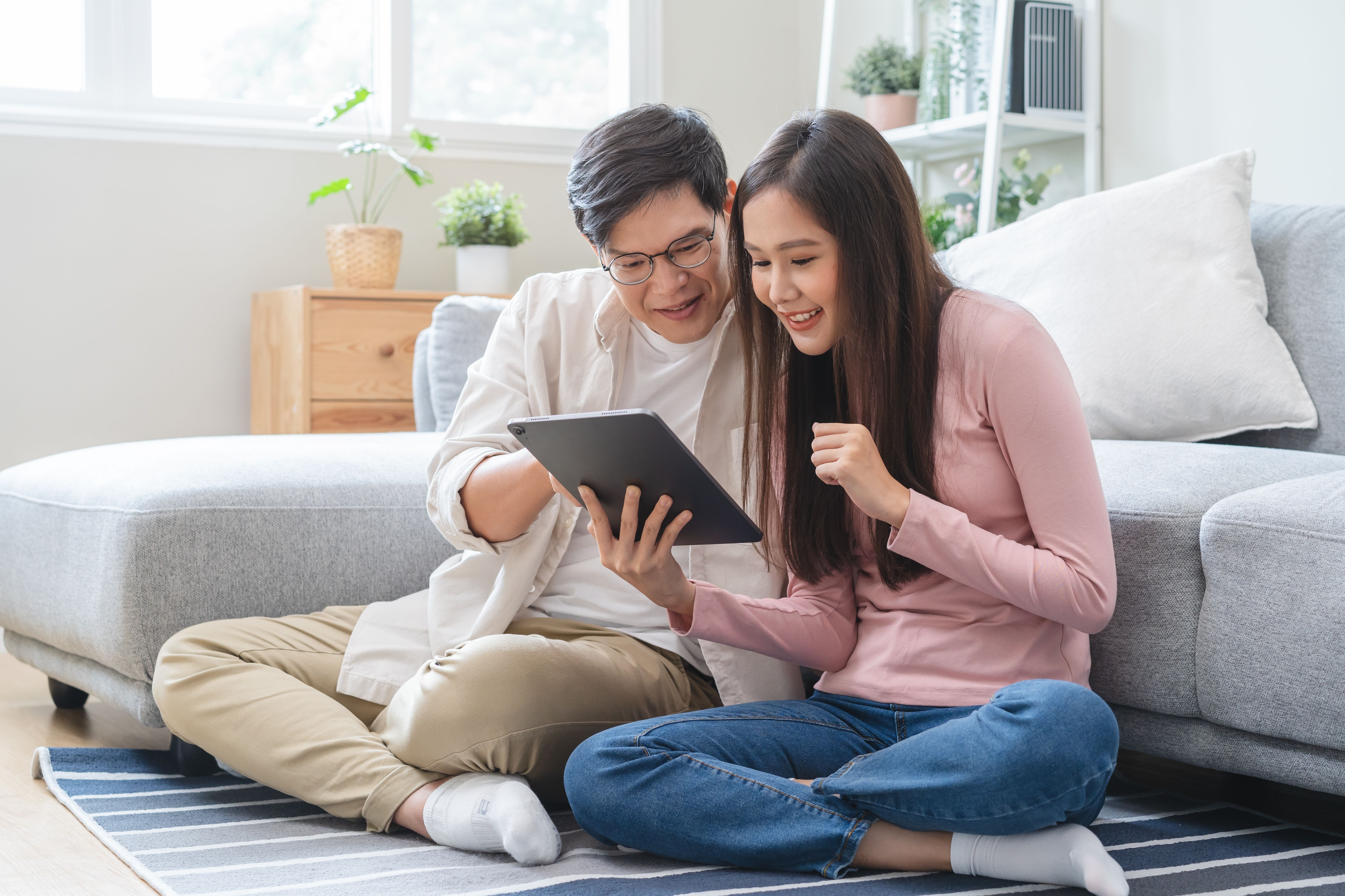 A young couple sitting on a rug in a living room, looking at a tablet and smiling together