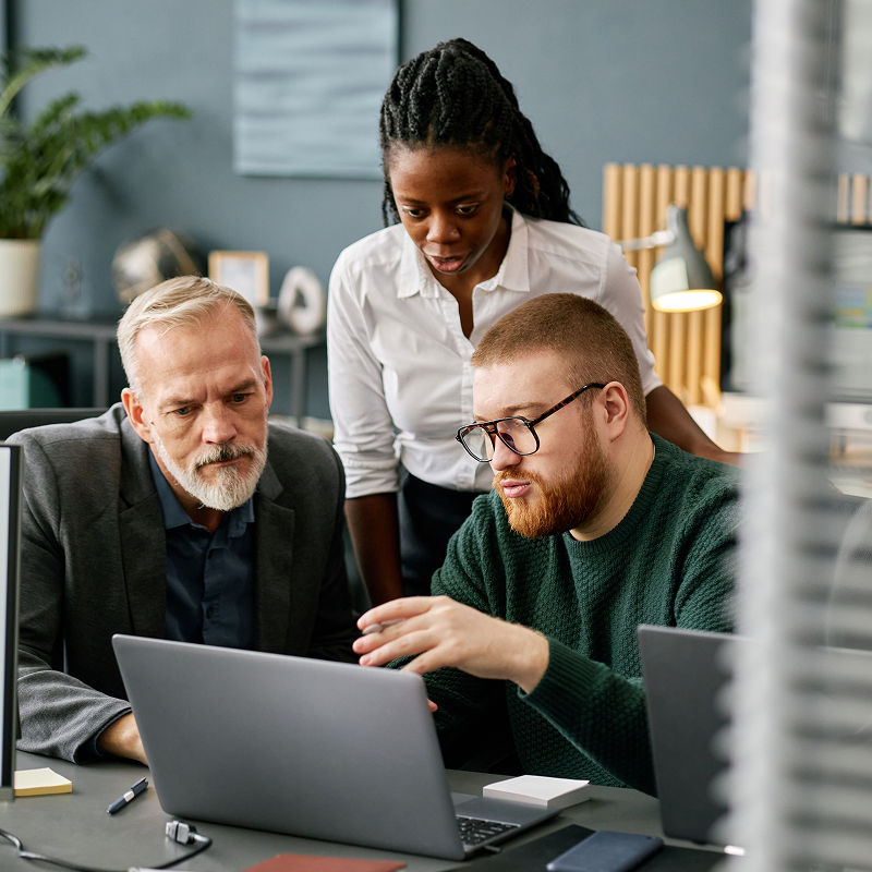 Two men sitting looking at a laptop with a woman standing behind them looking at the laptop screen