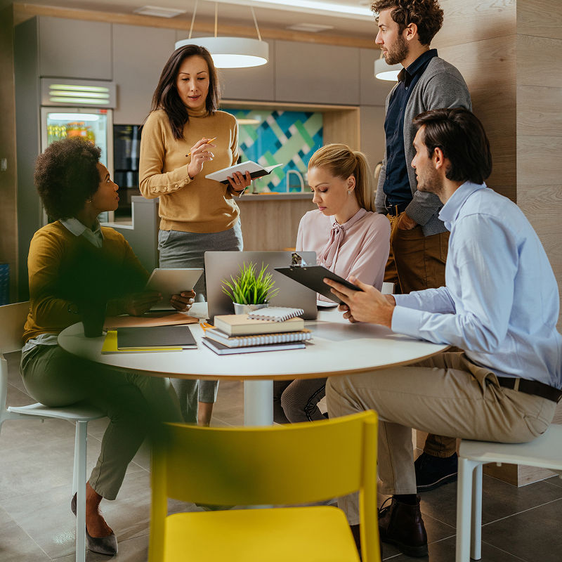 Three people sitting at a roundtable with tablets looking up at two others standing with tablets