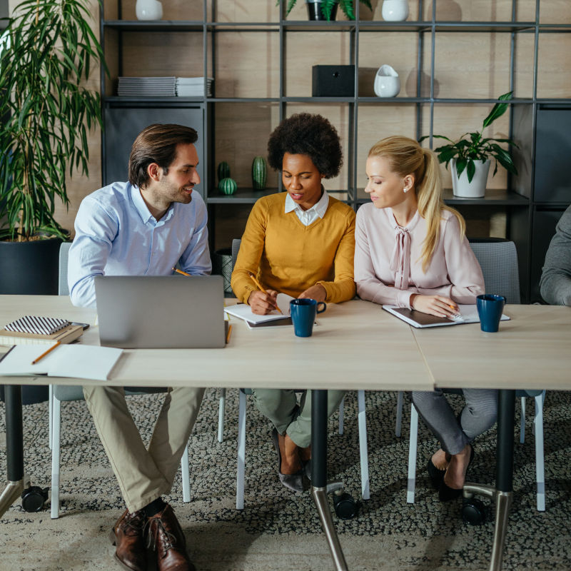 Three people sitting a table, looking at a laptop and writing on paper