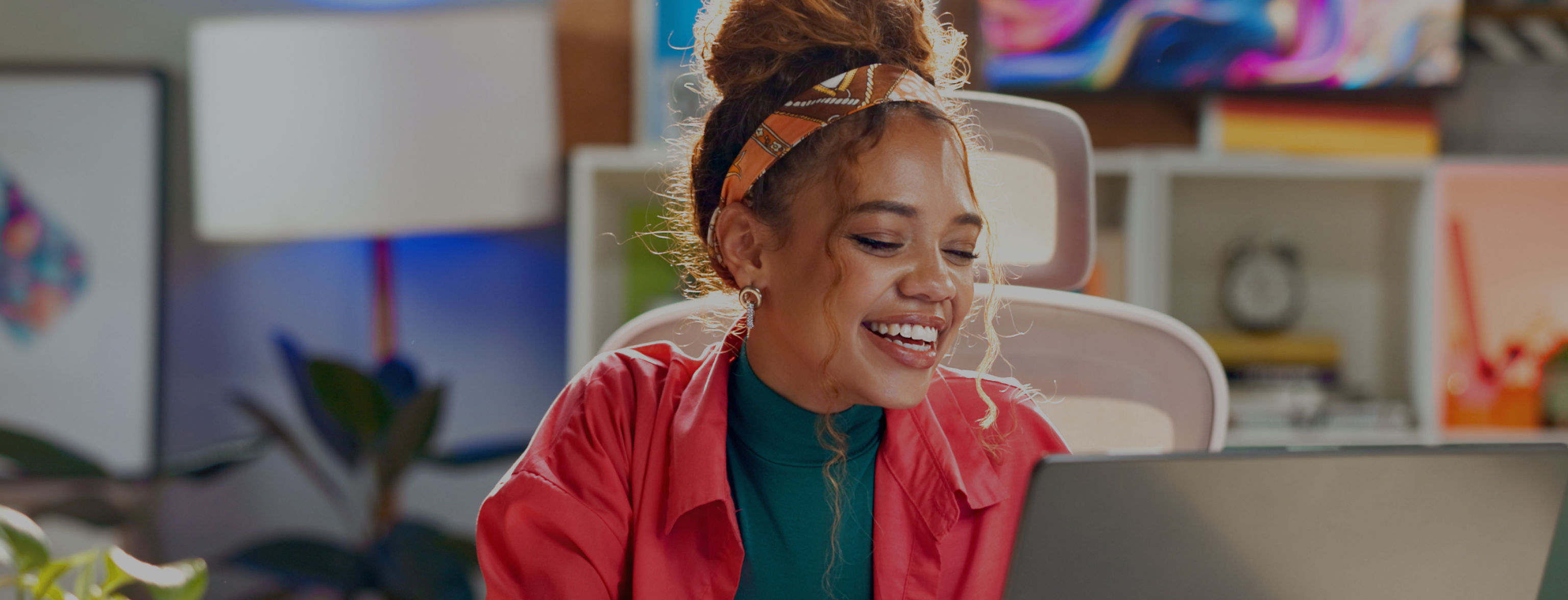 A woman dressed in a pink jacket and green top, laughing as she looks at a laptop screen
