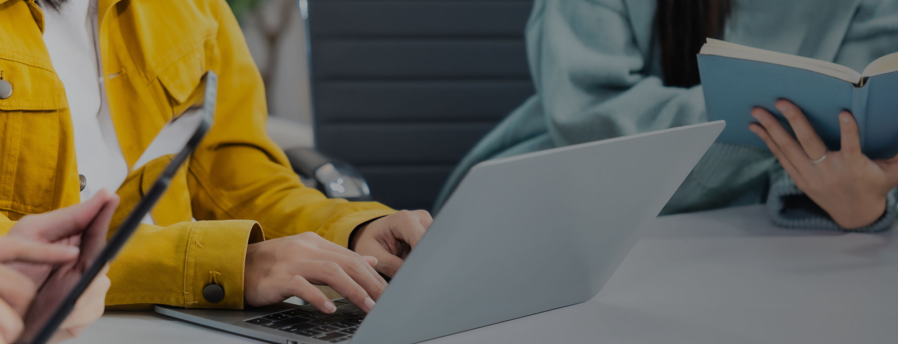 Drei Personen sitzen an einem Tisch: eine arbeitet am Laptop, eine hält einen Notizblock, eine ein Tablet.