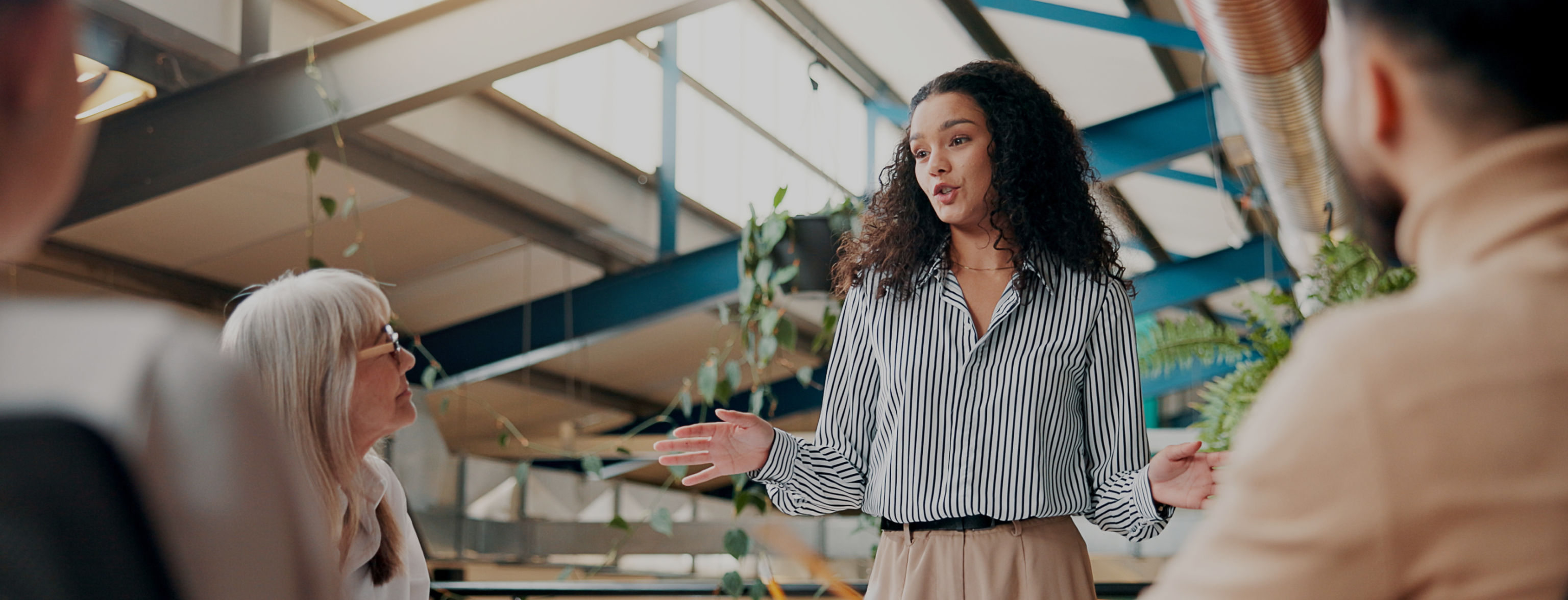 A woman standing in front of a group of people sat at a desk, arms outstretched explaining her ideas