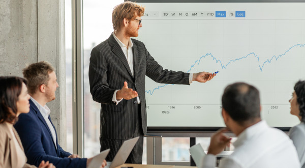 A man presenting to four other people, pointing at a line graph on a screen