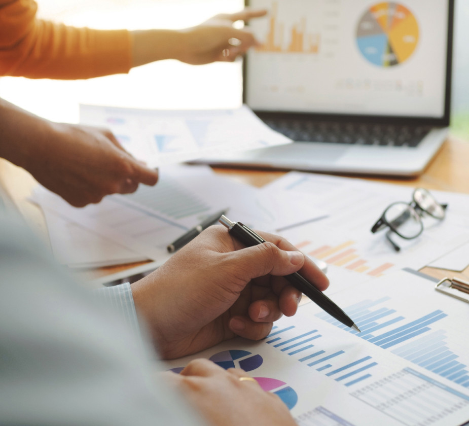 A figure pointing at a laptop screen  with paper and glasses sitting on a desk