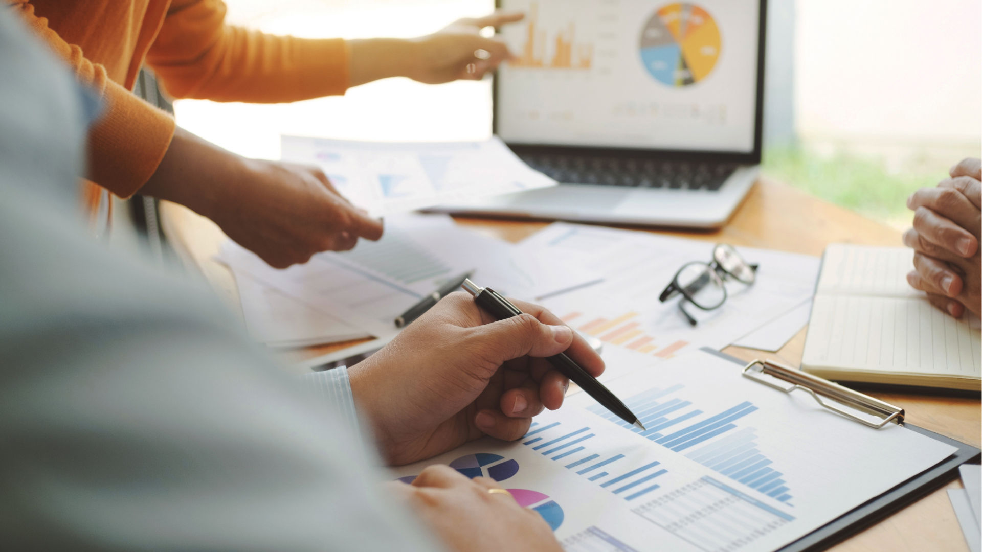 A figure pointing at a laptop screen  with paper and glasses sitting on a desk